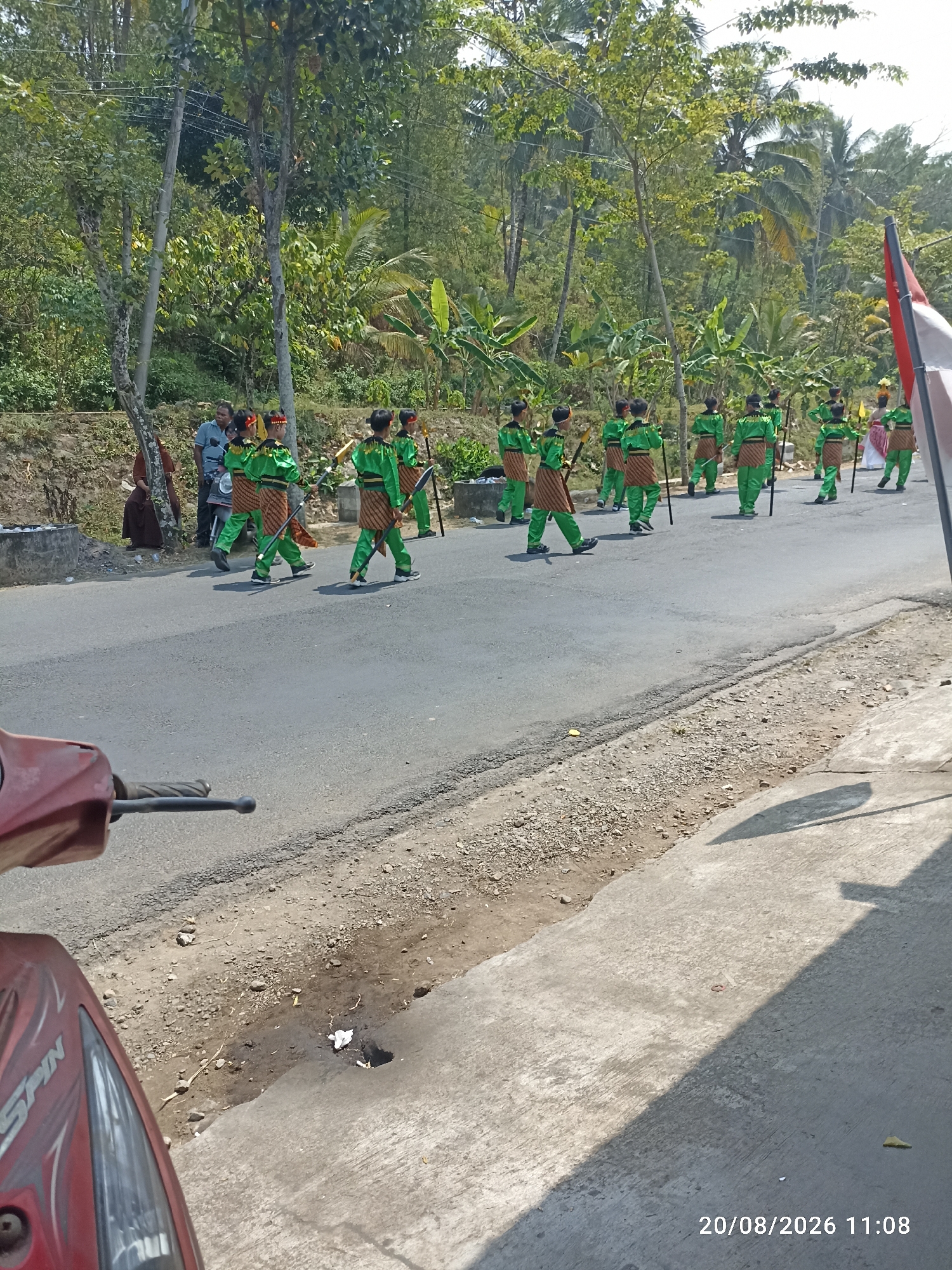In Munjungan, Indonesia, on August 20, 2026, at 11:08 AM, a group of young people dressed in green outfits and patterned sashes march in single file on an asphalt road. They carry sticks or poles. A few adults and a woman in a brown garment observe from the side of the road, near a stone wall and trees. The road is flanked by vegetation, including trees and palm trees, suggesting a rural or semi-rural environment. A portion of a red motorcycle is visible in the foreground on the left side of the image. The weather appears sunny.