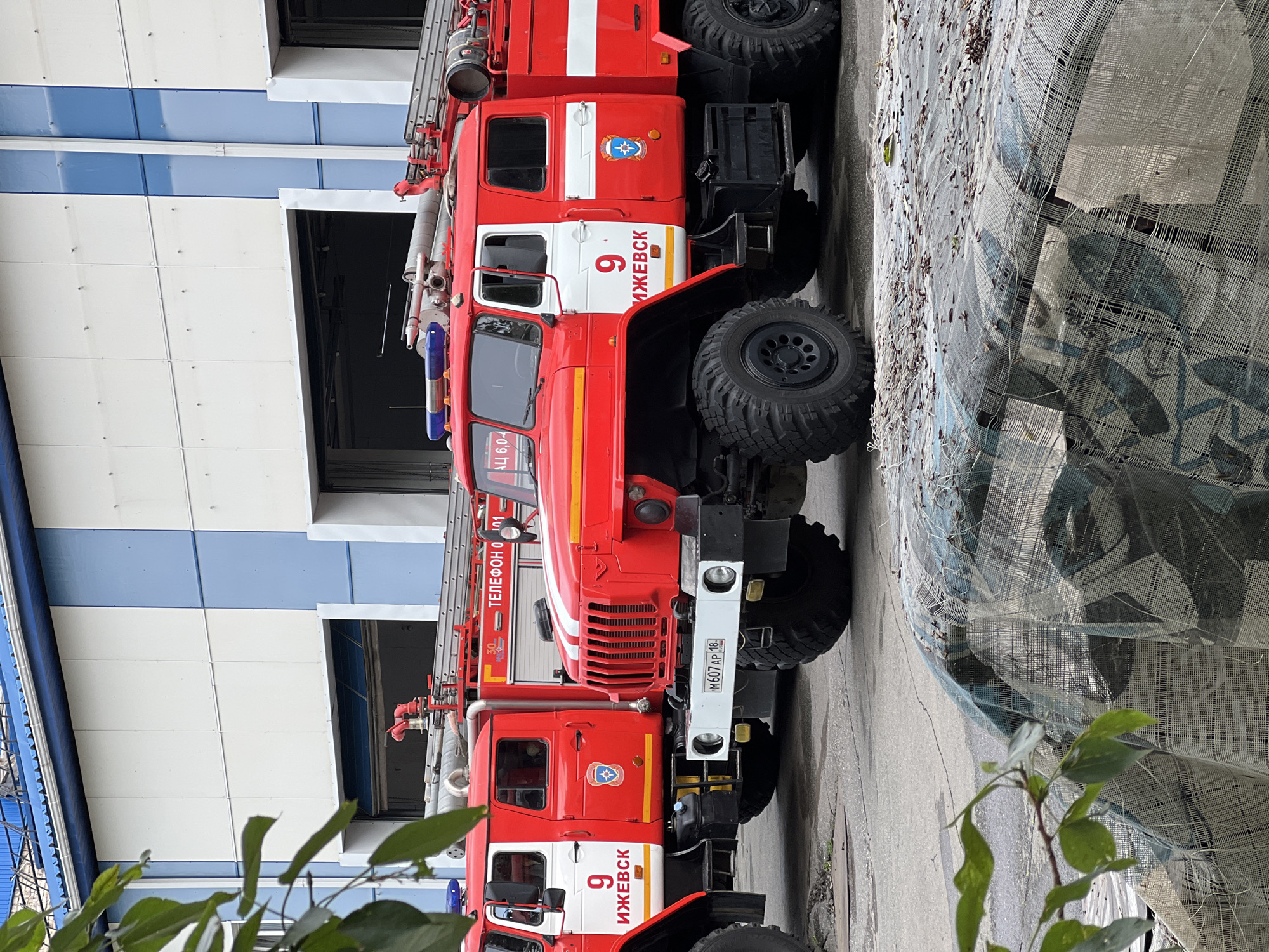 The image shows two red fire trucks parked outside a building with blue and white panels. The fire truck in the foreground has the number "9" and "ИЖЕВСК" (Izhevsk) written on its side. The license plate on the front bumper reads "M607AP 18". A covered object, possibly a tarp, is in the foreground, with some green leaves hanging from the left side of the frame. The building appears to be a fire station or garage, with large open doorways visible. The scene is set in the city of Izhevsk, Russia.