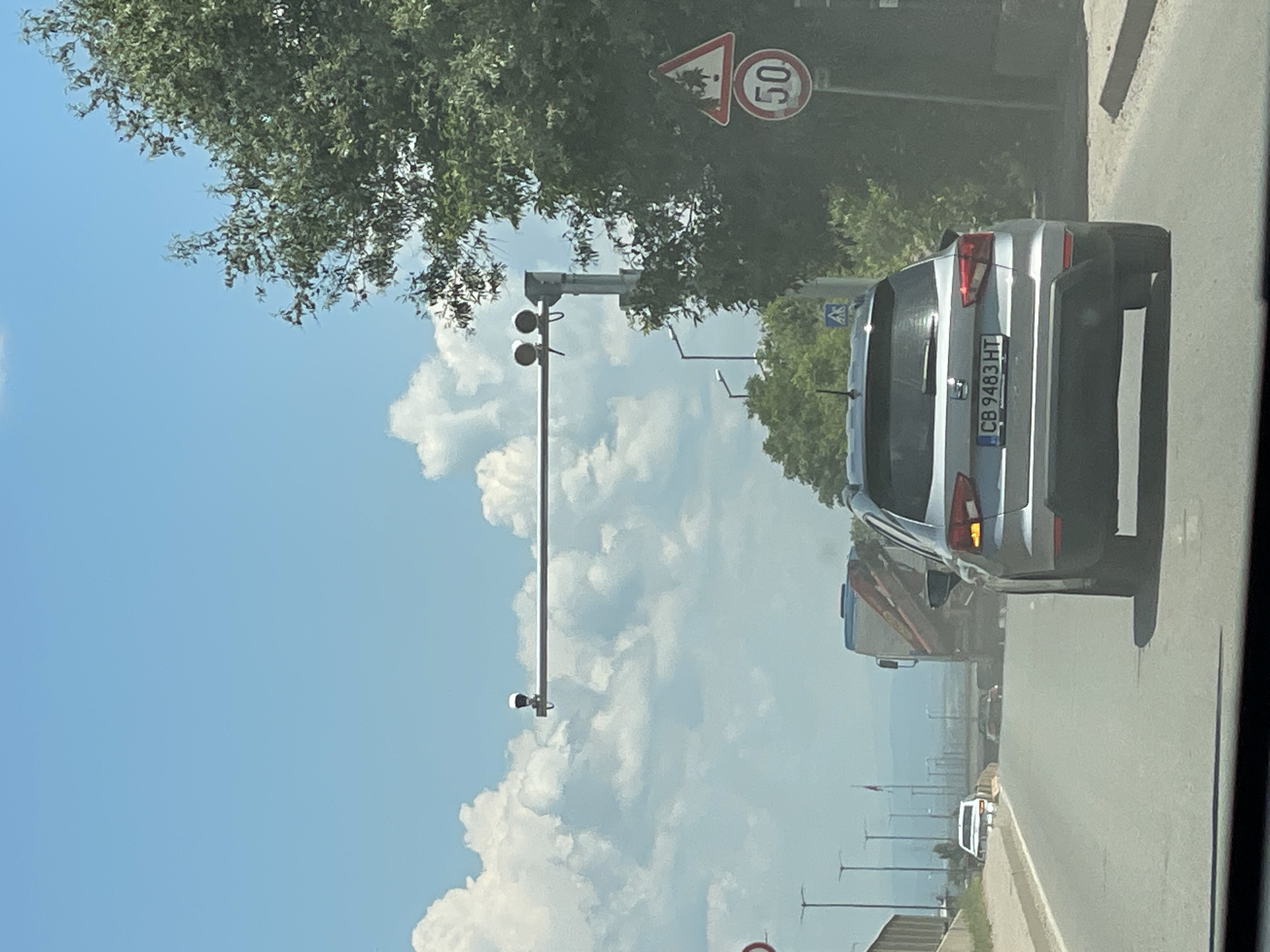 The image shows a view from a vehicle driving on a road in Sofia, Bulgaria. A silver SUV with license plate CB 9483 HT is in the foreground. In the background, there is a truck with a crane, other vehicles, traffic lights, street lights, trees, and a speed limit sign indicating 50. The sky is blue with white clouds.