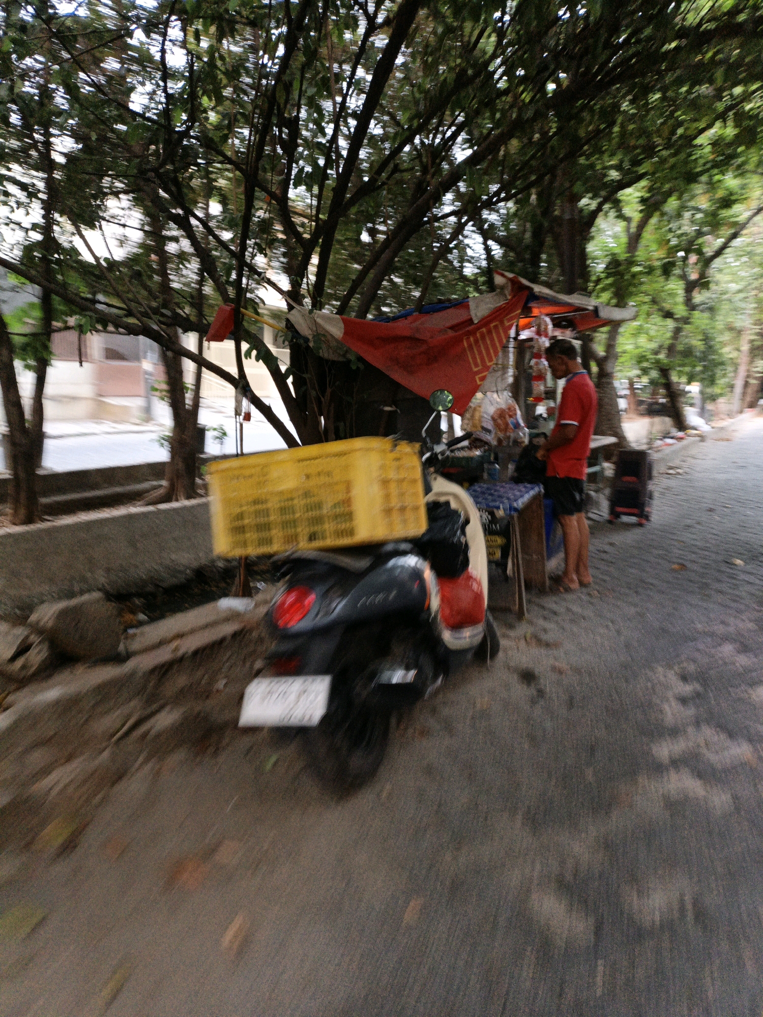 A man in a red shirt and black shorts stands in front of a food stall under a canopy of trees, in what appears to be a street in North Jakarta, Indonesia. The food stall is set up with a red tarp and a blue and white patterned tablecloth on a wooden stand. Behind the man, a yellow plastic crate is attached to a motorcycle. The motorcycle is partially visible, with a red taillight and a white license plate. The scene is cluttered with bags and other items associated with a street food vendor. The ground is paved with asphalt, and there is a concrete barrier to the left of the stall. The image is slightly blurred, suggesting movement.
