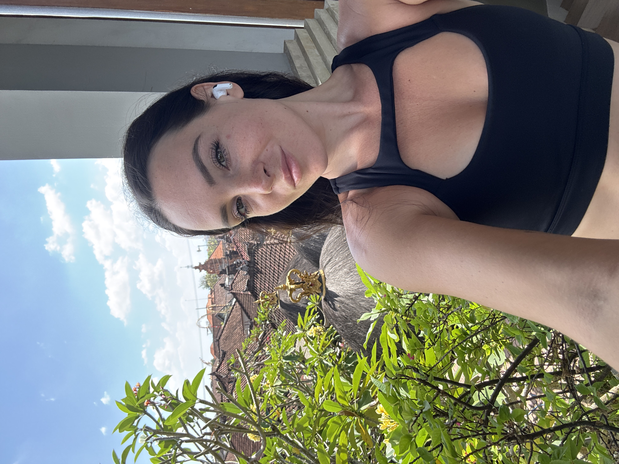 In North Kuta, Indonesia, a woman with dark hair and fair skin takes a selfie outdoors. She is wearing a black sports bra with a cutout on the chest and white wireless earbuds. Her expression is relaxed and she is looking towards the camera. In the background, there is lush green foliage with yellow flowers, red-tiled roofs with ornate golden crowns on top, and a bright blue sky with scattered clouds. A staircase is visible to the right of the woman.