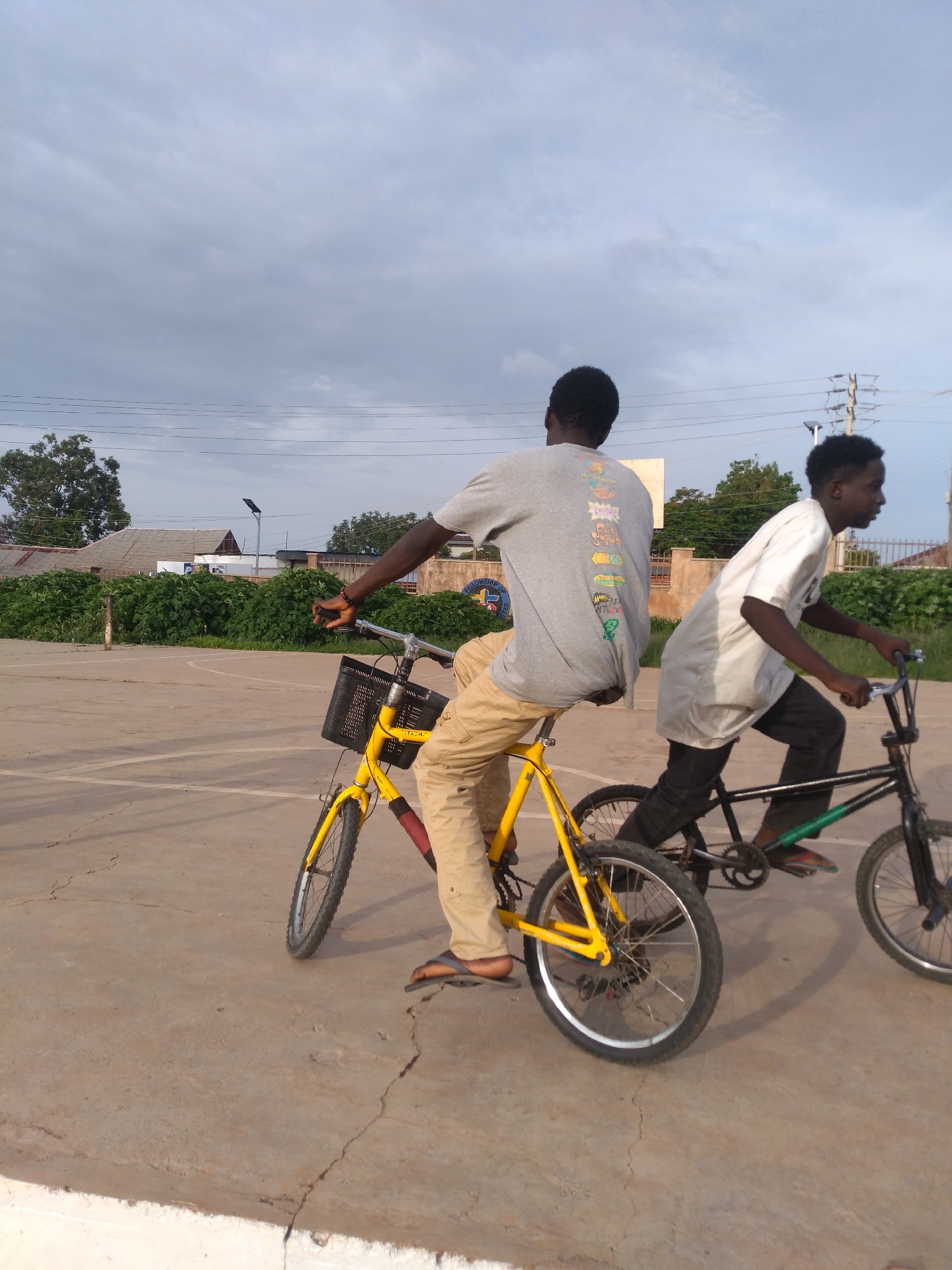 The image depicts two young men on bicycles in an outdoor urban setting. The person in the foreground is riding a yellow bicycle with a black basket attached to the handlebars. They are wearing a grey t-shirt with a graphic design and tan pants. The second person is on a black bicycle, wearing a white t-shirt and black pants. Both individuals appear to be moving forward on a paved surface that resembles a court or parking lot, with cracks visible in the pavement. In the background, there are buildings, dense green vegetation, and utility poles with power lines. The sky is overcast with grey clouds. The scene appears to be in the city of Unknown, Unknown.