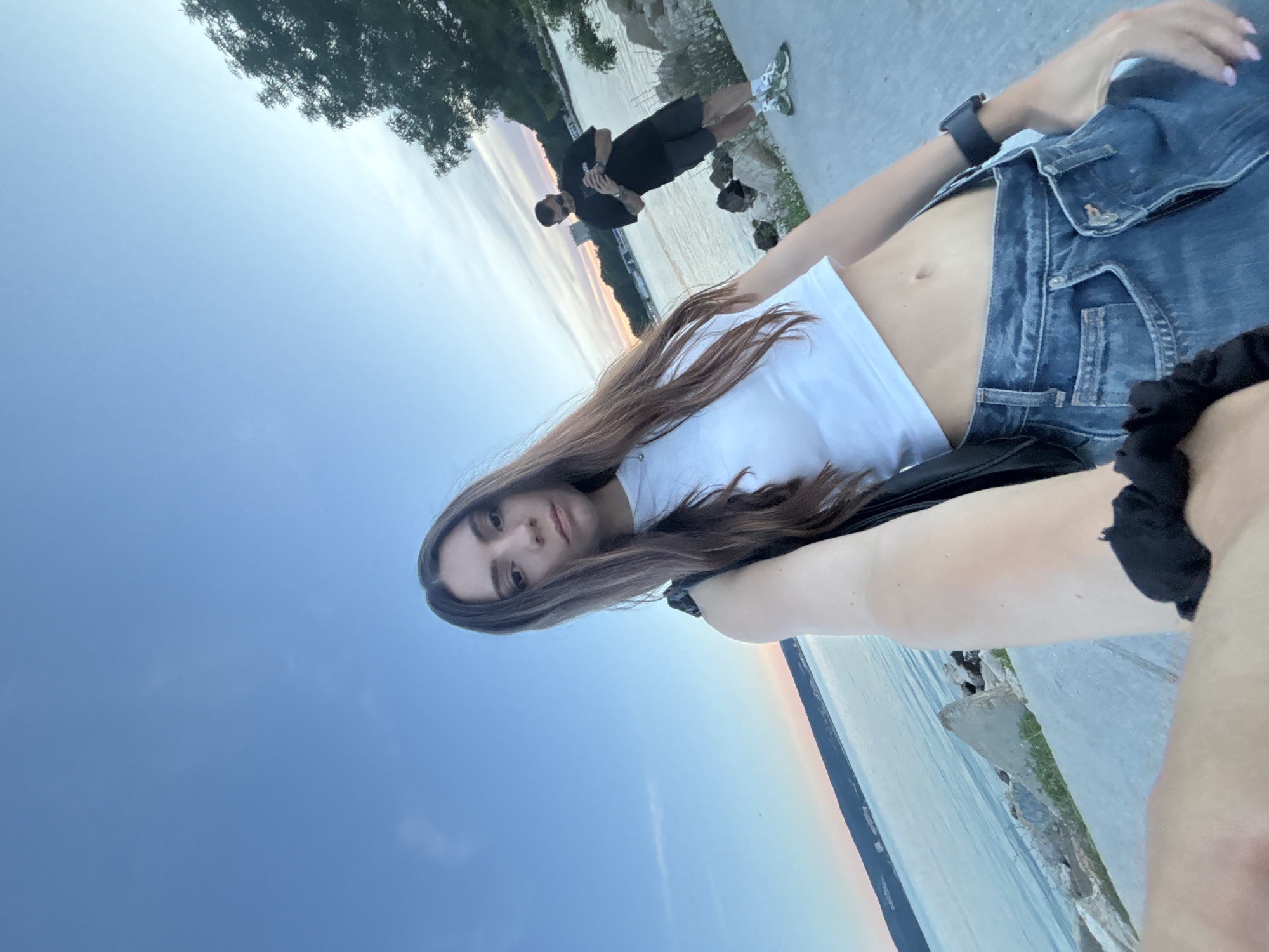 A woman is taking a selfie outdoors with a body of water in the background. She is wearing a white crop top and denim jeans. Behind her, a man is standing and looking at his phone. The scene appears to be a waterfront promenade, possibly in Varna, Bulgaria, given the presence of a coastal landscape and a generally clear sky transitioning into twilight. Rocks and vegetation are visible along the water's edge. The overall lighting suggests it is late afternoon or early evening.