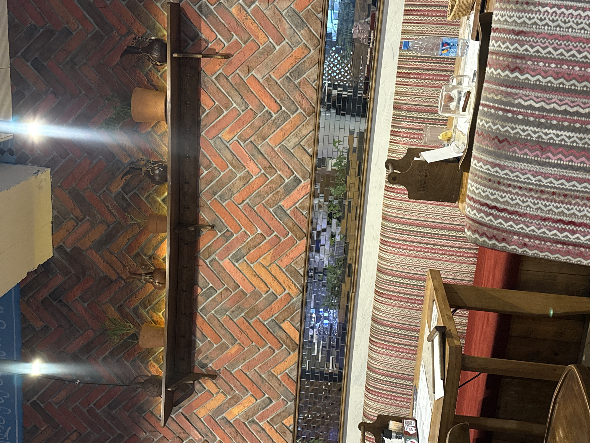 The image depicts the interior of a restaurant in Alushta, Ukraine, with a focus on a brick-patterned wall and seating area. The wall, constructed with small, diagonally laid bricks in shades of orange and red, features a wooden shelf displaying several small potted plants and decorative objects. Below the shelf, a long, narrow mirror reflects the room, showing hints of greenery and possibly other decorative elements. In the foreground, a wooden table is set with placemats, glasses, and a bottle of water. Behind the table, a banquette seat is covered in a striped fabric in red, white, and pink. A portion of another chair with a patterned fabric is visible on the right side of the frame. The lighting suggests an indoor setting, possibly during evening hours. There are no people visible in the image.