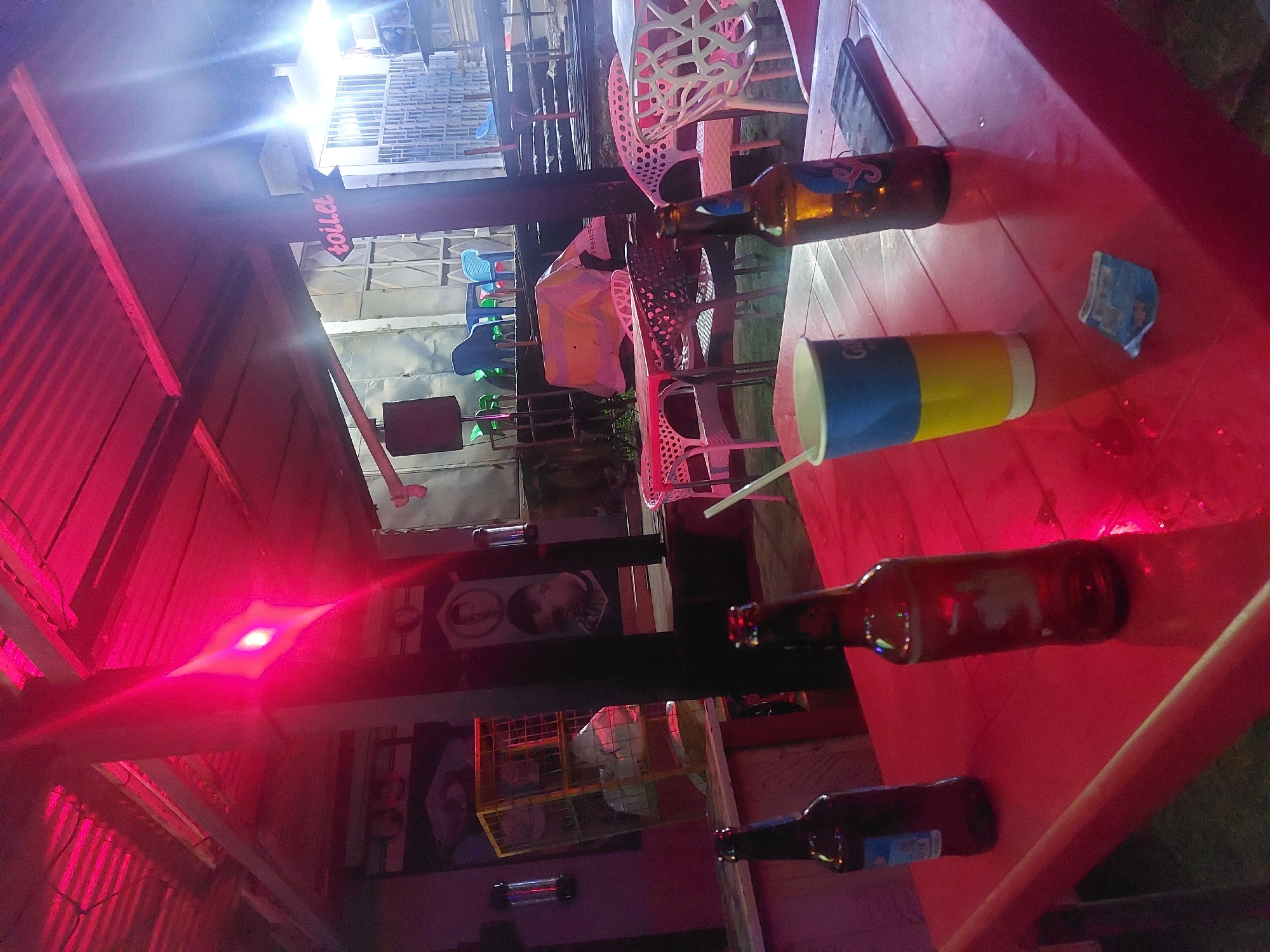 The image depicts a nighttime scene, likely outdoors, at a bar or restaurant in Dar es-Salaam, Tanzania. The viewpoint is close to a red table.
Objects on the table include three beer bottles (brown, glass), a cup with blue, yellow, and white stripes, and a smartphone. There is also what appears to be a discarded wrapper or receipt. A straw is visible near the cup.
There are plastic chairs with ornate, open-weave backs situated around the table. The scene is lit with red light. In the background, there are buildings, a "toilet" sign, and various decorations on the walls, including a poster or picture of what appears to be a person's face. The overall impression is a casual, social setting.
