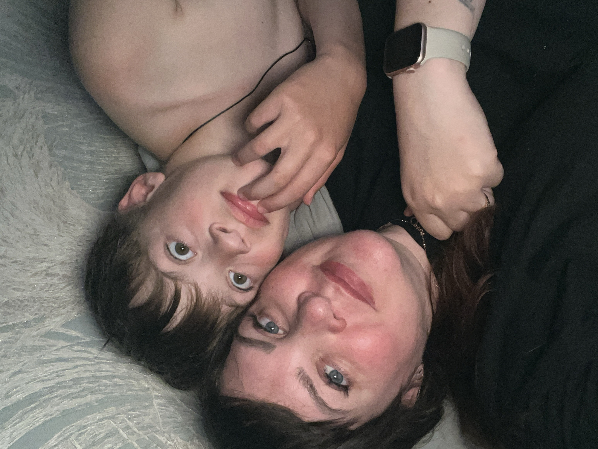 An overhead close-up shot captures an adult woman and a child lying down together, looking towards the camera. The scene takes place indoors, likely in a bedroom or living area in Noviy, Russia, given the comfortable, textured bedding.
The adult woman, positioned in the foreground and to the right, has dark hair, blue eyes, and a relaxed, gentle expression with a slight smile. She is wearing a dark-colored top. A smartwatch with a light-colored band and a dark screen with a rose gold-toned bezel is visible on her left wrist. A ring is also visible on one of her fingers. Her hair appears dark brown or auburn.
Next to her, the child, likely a boy, is positioned slightly above and to the left. He has short, dark brown hair and light brown or hazel eyes, looking intently at the camera with a soft expression. He is shirtless. His left arm is draped over the woman's upper body, with his fingers gently touching near her chin and mouth, suggesting affection or playfulness.
They are lying on a soft, light-colored, textured fabric, possibly a duvet or blanket, which features a subtle pattern. The overall lighting is soft, creating an intimate and peaceful mood. There is no visible text in the image.