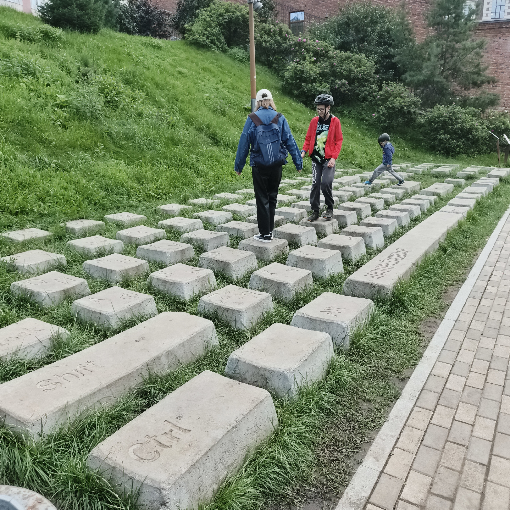 Three people, two teenagers and a younger child, are walking across a path made of large concrete blocks shaped like computer keys. The teenagers are wearing helmets; one is wearing a denim jacket and a backpack, the other a red jacket. The child is wearing a blue hoodie. The scene is outdoors, with a grassy hillside behind the path. The concrete blocks are arranged in rows, mimicking a keyboard layout. Some of the blocks have visible text etched into them, including "Ctrl", "Shift", and what appears to be "Lock". The setting seems to be a park or public space in Yekaterinburg, Russia, indicated by the location context. The weather appears to be overcast or slightly cloudy, and the time of day is daytime. The overall mood is casual and recreational, with the individuals enjoying the unique pathway.