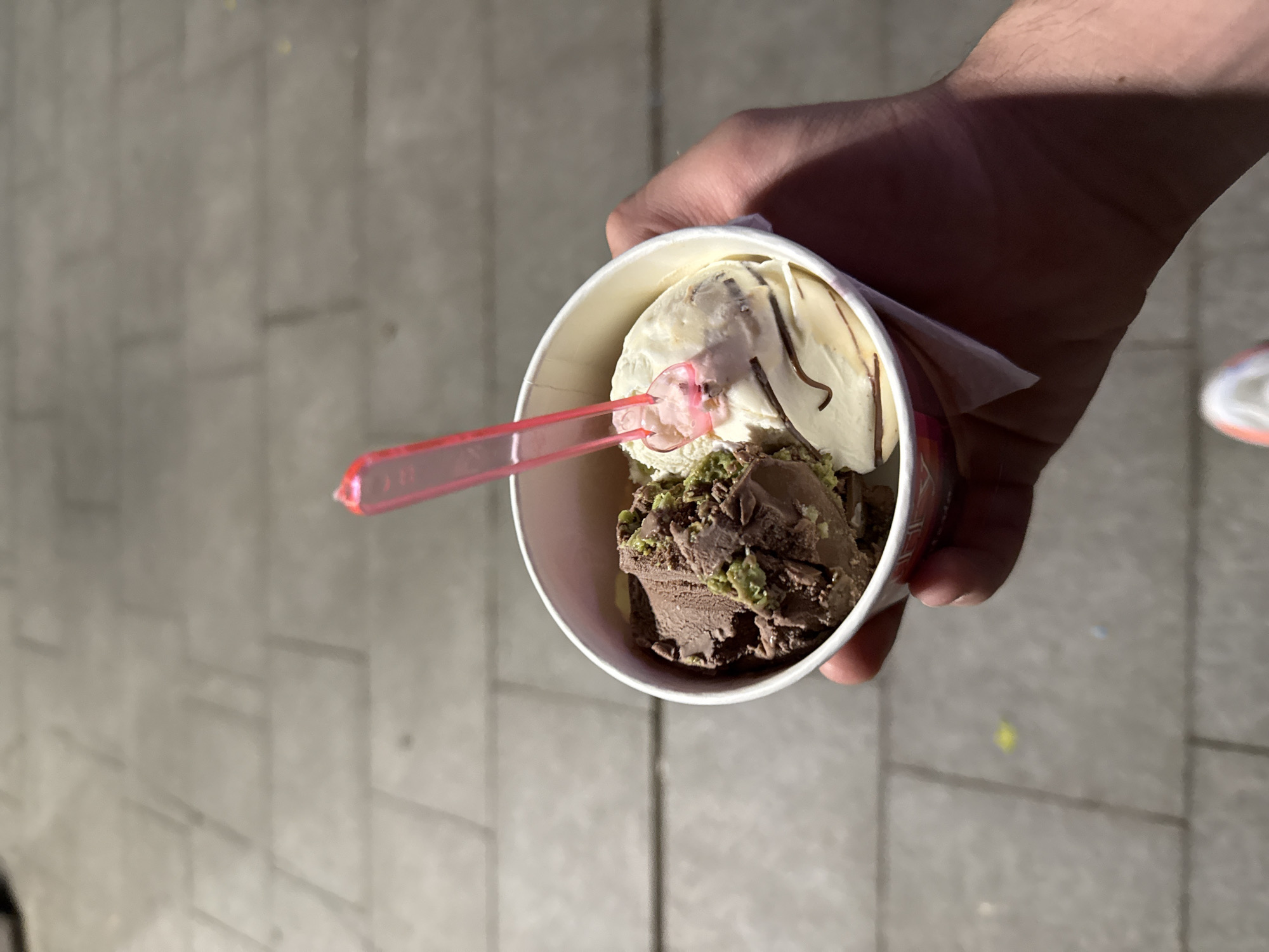 A hand is holding a paper cup with two scoops of ice cream, one chocolate with green pistachios and the other vanilla with chocolate swirls, and a pink plastic spoon inserted in it, on a sidewalk in Stara Zagora, Bulgaria. The sidewalk is paved with grey rectangular tiles.
