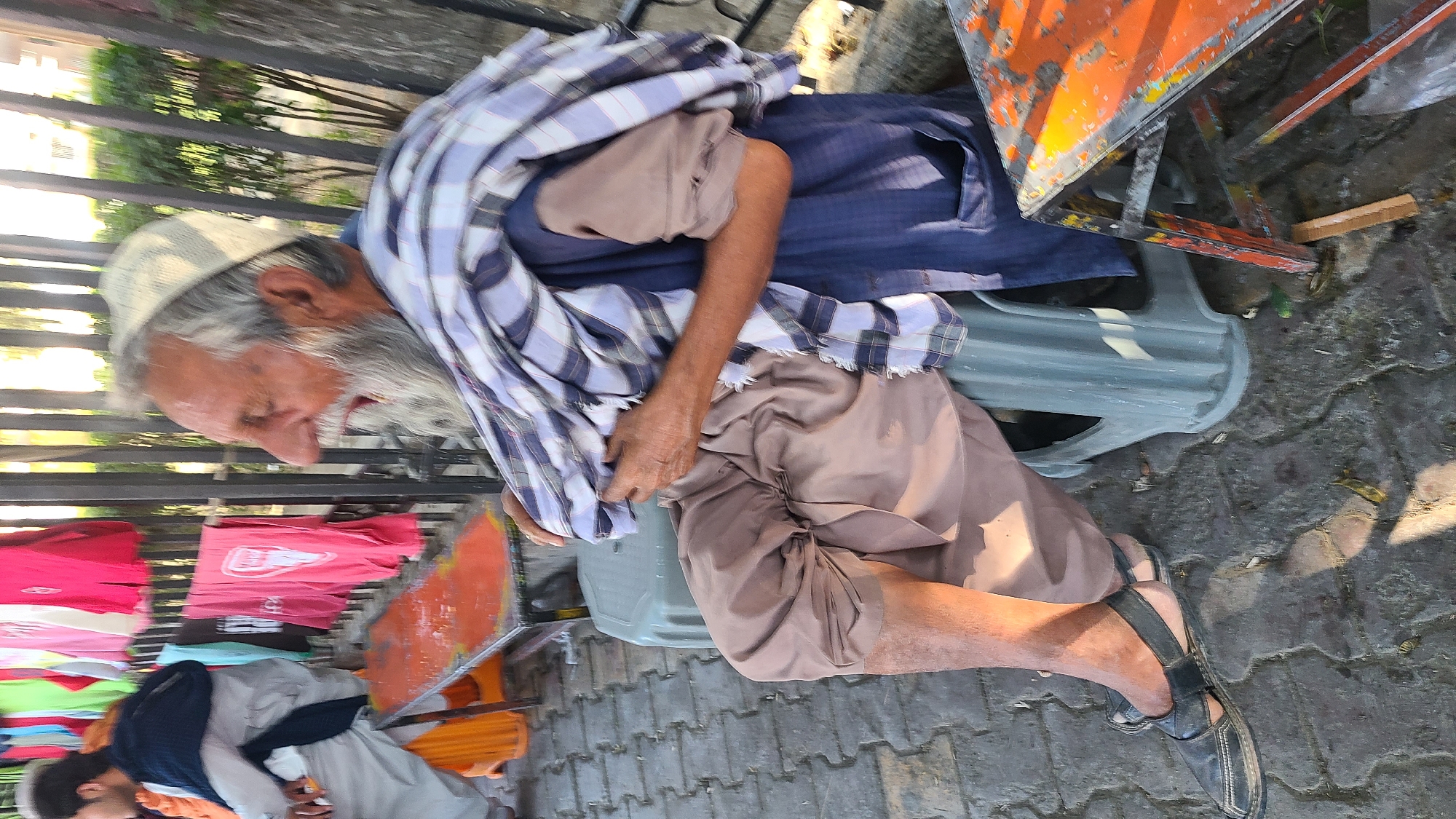 A senior man is seated on a plastic chair outdoors. He has a long white beard, wears a white cap, and is draped in a blue and white plaid shawl. He is wearing a blue shirt and brown pants, and black sandals on his feet. Next to him is a metal rack with colorful clothes hanging on it. An orange metal object is next to him. The ground is paved. The image suggests the location is in a city in Pakistan.