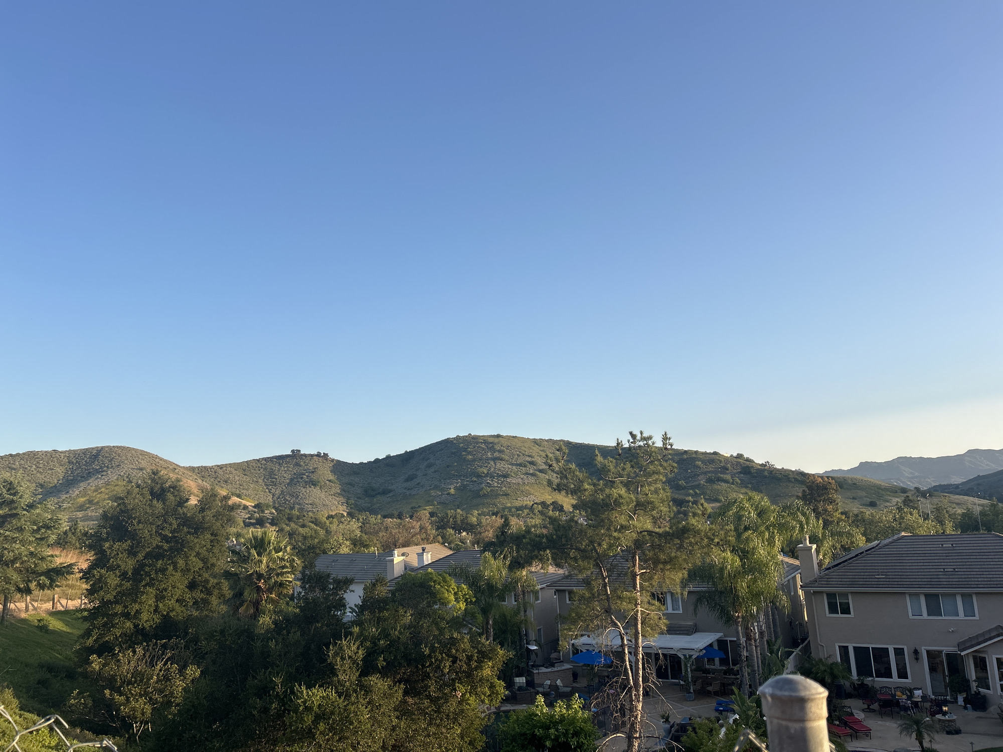 The image depicts a daytime view in Oak Park, United States of America, overlooking residential properties and rolling hills. The sky is clear blue. Several houses with tan exteriors and grey tiled roofs are visible among trees, obscuring the full view. Hills covered in vegetation form the backdrop. The immediate foreground shows foliage and a partial view of a fence.