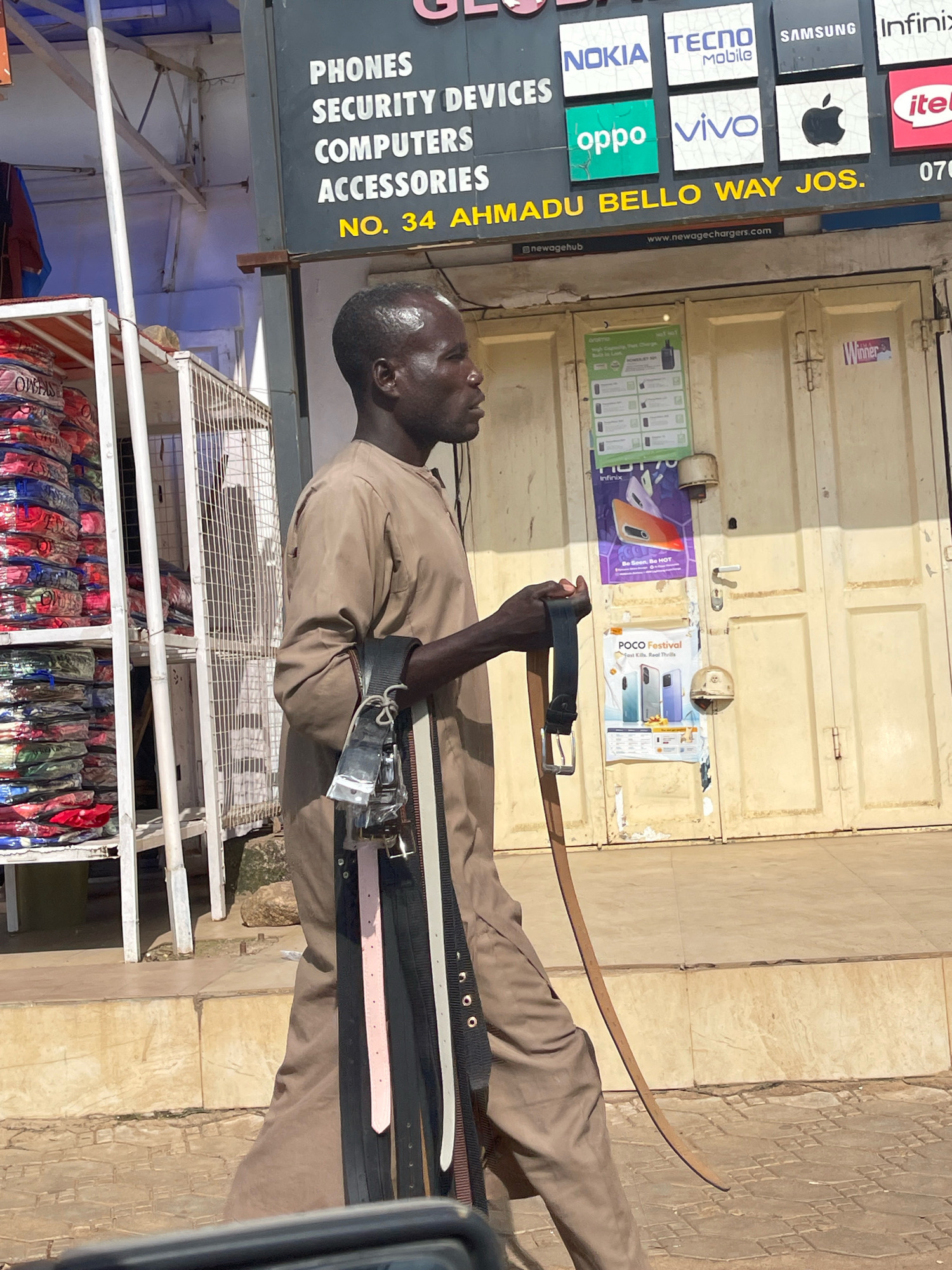A man in a tan robe walks along a paved sidewalk in Jos, Nigeria. He is carrying several belts draped over his left arm and shoulder. Behind him, a storefront displays signs for mobile phone brands like Nokia, Tecno, Samsung, Infinix, Oppo, and Vivo, as well as an Apple logo. A sign above the entrance reads "PHONES SECURITY DEVICES COMPUTERS ACCESSORIES NO. 34 AHMADU BELLO WAY JOS." To the left of the storefront, racks of colorful packaged clothing are visible. The man's right arm is extended, holding a curved object that resembles a scimitar. The scene is lit by natural daylight.