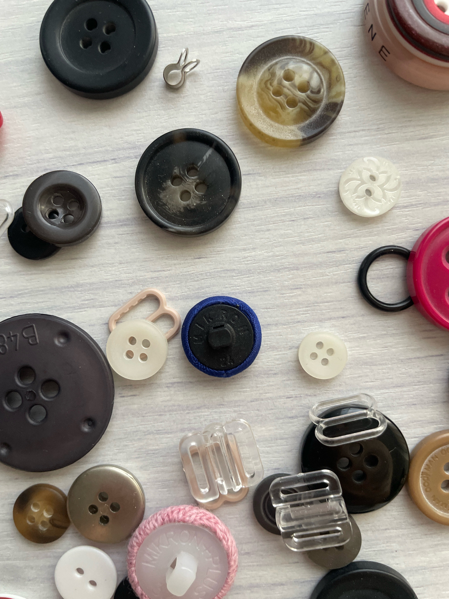 The image is a close-up, top-down view of a collection of assorted buttons and sewing accessories scattered on a light-colored, wooden surface. The buttons vary in size, color, material, and design, including classic four-hole buttons, decorative buttons with intricate patterns, and fabric-covered buttons. There are also a few plastic clasps and a small metal loop. The lighting is bright and even, suggesting an indoor setting. There is no indication of the city of Atarfe, Spain, or any people or actions depicted in the image.