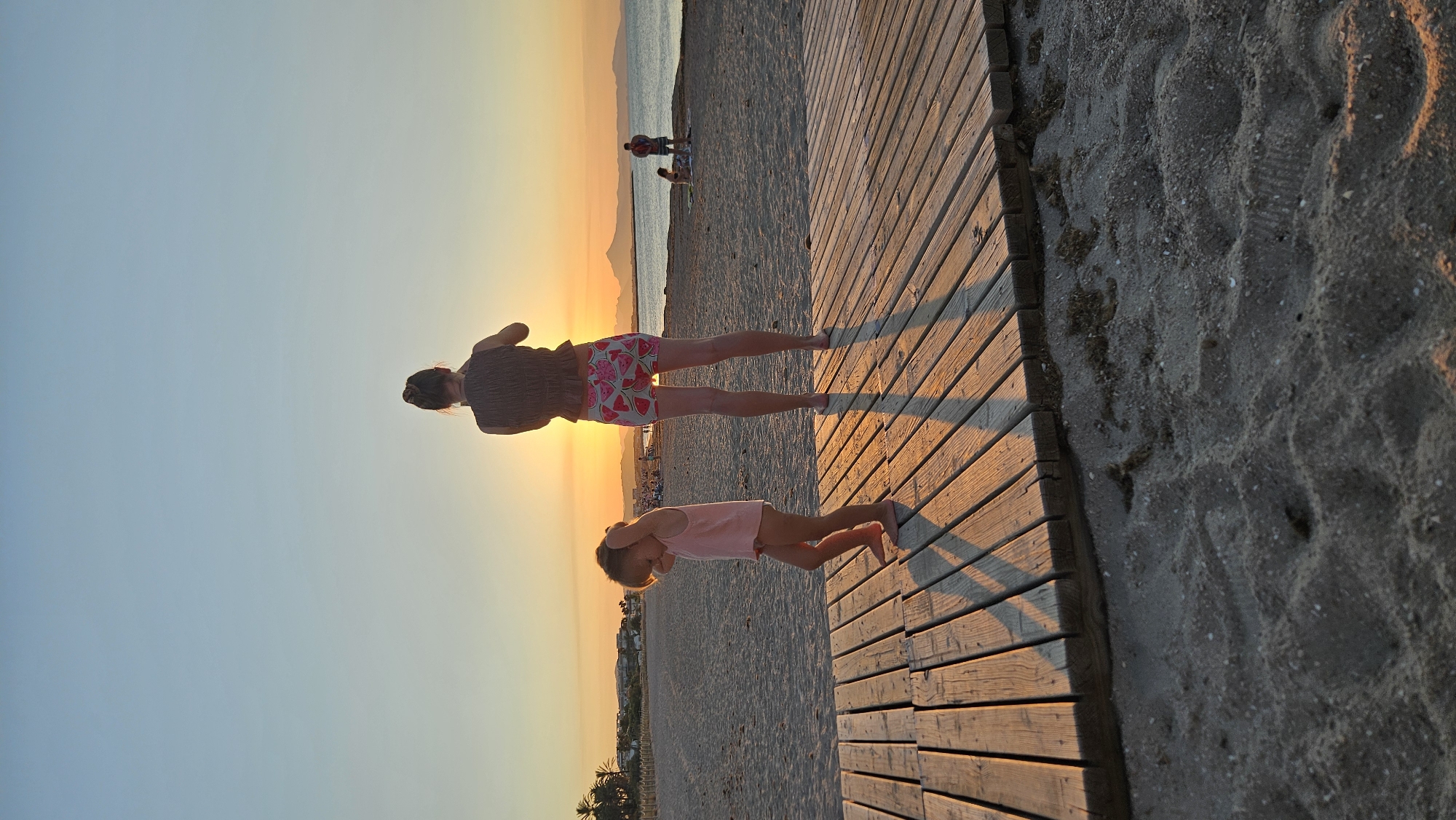Two individuals, a woman and a young child, stand on a wooden boardwalk at the beach in Deveses, Spain, during sunset. The woman, viewed from behind, wears a dark, textured top and shorts with a watermelon pattern. She is holding up what appears to be a phone, possibly taking a picture. The child, also viewed from behind, is wearing a pink tank top and a diaper or similar undergarment. The child has their hands on their head and appears to be looking down. The boardwalk is constructed of wooden planks and extends across the sand. In the background, the sun is setting over the sea, casting a warm orange glow across the sky and the beach. Other beachgoers are visible in the distance, as well as palm trees and buildings along the coastline. The sand is textured with footprints and scattered debris.