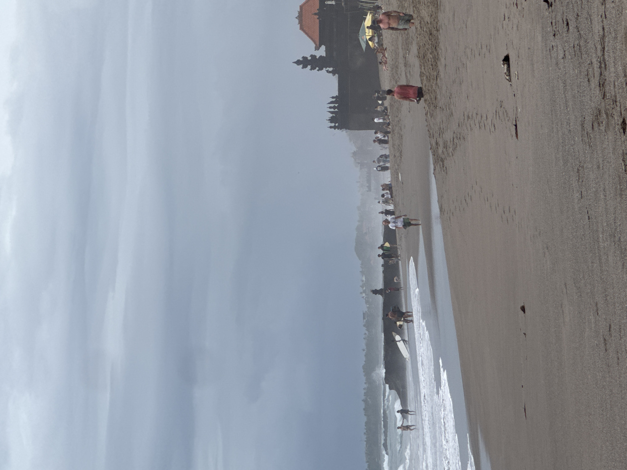 The image displays an active beach scene in North Kuta, Indonesia, under an overcast sky. The primary subjects are numerous people scattered along a wide, sandy shoreline and in the shallow ocean waters. In the mid-ground, a dark, ornate building with a distinctive reddish-brown tiled roof, characteristic of Balinese temple architecture, stands prominently, overlooking the beach.
People are engaged in various beach activities: many are walking along the water's edge, some are standing and observing the ocean, and others appear to be relaxing. Several individuals are visible with surfboards near the breaking waves, indicating surfing or paddleboarding activities. A yellow beach umbrella offers shade near the temple-like structure. The sky is a uniform light grey, suggesting an overcast day or possibly a hazy atmosphere, giving the scene a soft, diffused light. The beach sand is a light tan, and white foam marks the breaking waves close to the shore. No visible text is present in the image.