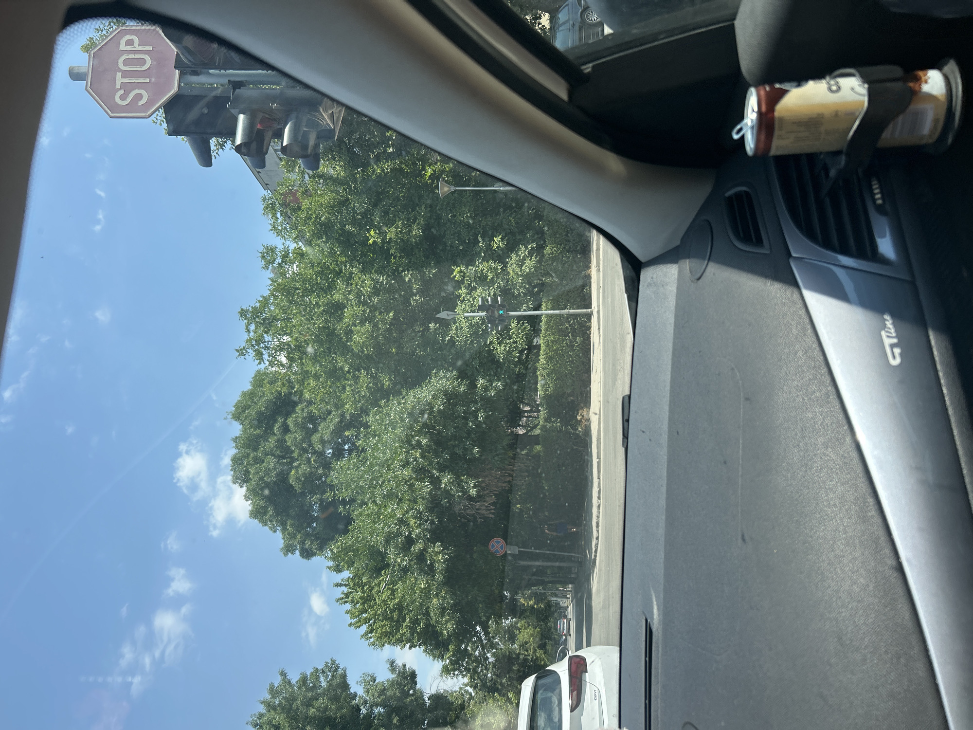 The image shows a view from inside a car looking out towards a street intersection on a sunny day. The car's dashboard and a portion of the passenger side are visible in the foreground, with a can of drink in a cup holder. Outside the car, there is a traffic light with a stop sign above it. Lush green trees line the street, and several cars are visible in the distance. A person can be seen walking on the sidewalk. The scene appears to be a typical urban environment. Based on visual cues such as architecture and vegetation, this location is likely Plovdiv, Bulgaria.