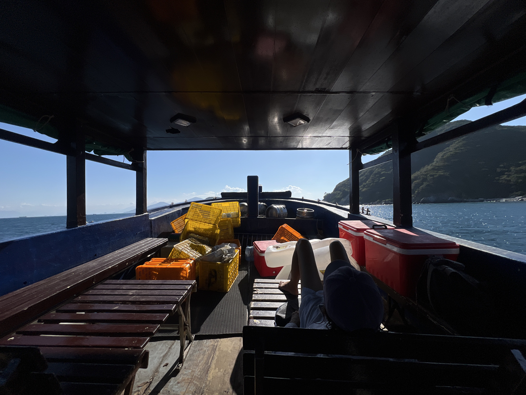 The image shows the interior of a boat, likely a fishing boat, on the water. The location is suggested to be near Bac Nha Trang Ward, Vietnam, based on the sea and coastal geography. The boat is filled with yellow and orange crates, two red coolers, and other containers. A person is reclining on a wooden bench towards the rear of the boat. The environment consists of the sea, sky, and mountainous coastline visible in the background through the boat's open sides. The deck is flat in the middle and the benches line the side of the boat. The overhead is covered with dark wood. There are no distinct actions or interactions visible in the image.