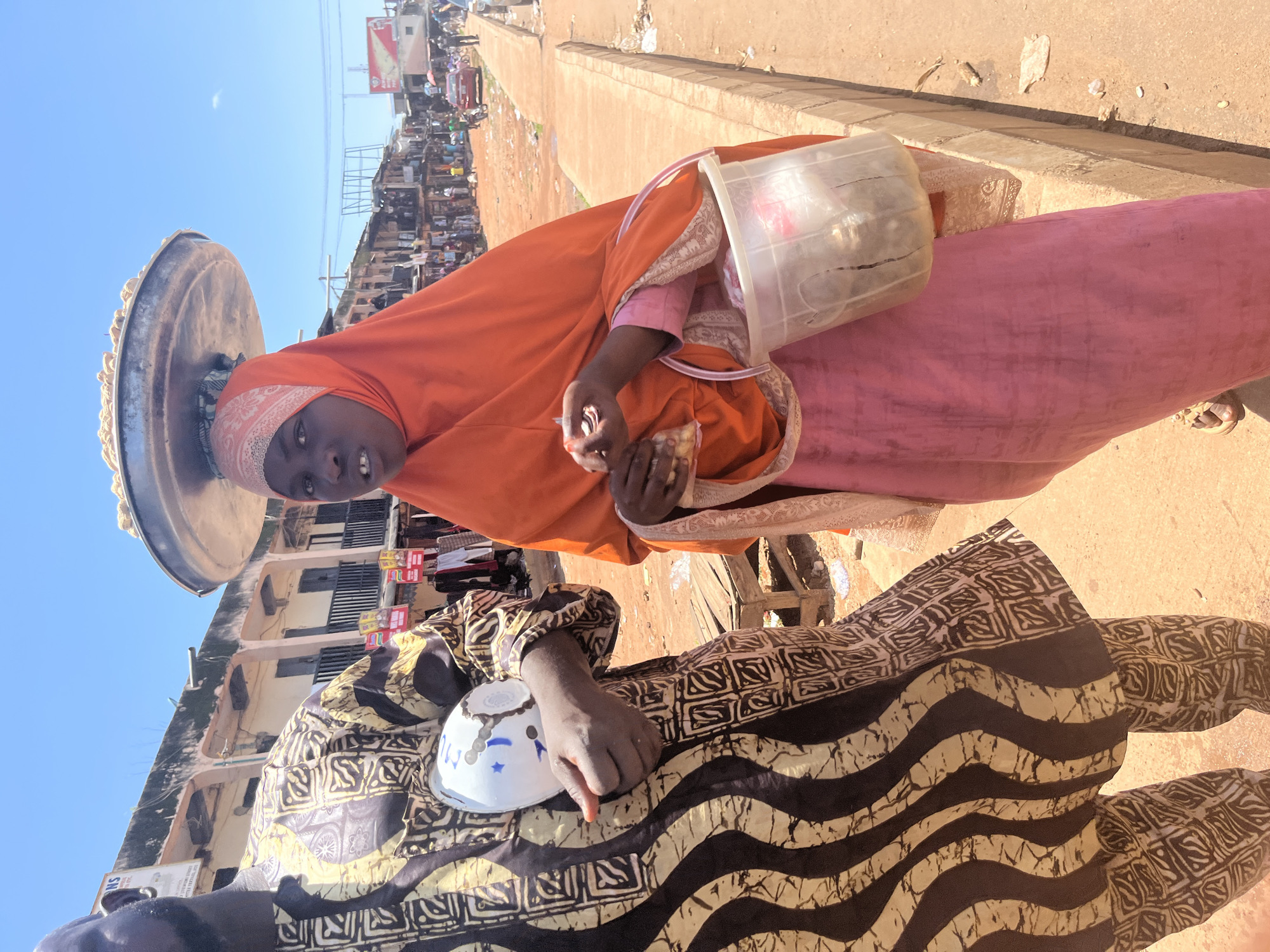 In Jos, Nigeria, a woman wearing an orange hijab and a pink dress balances a large metal tray of food on her head. She holds a small bag of items and a clear plastic bucket filled with food in her right hand. Beside her, a man in a black and gold patterned robe holds a white and blue object. The scene takes place on a dusty street with buildings and market stalls in the background.