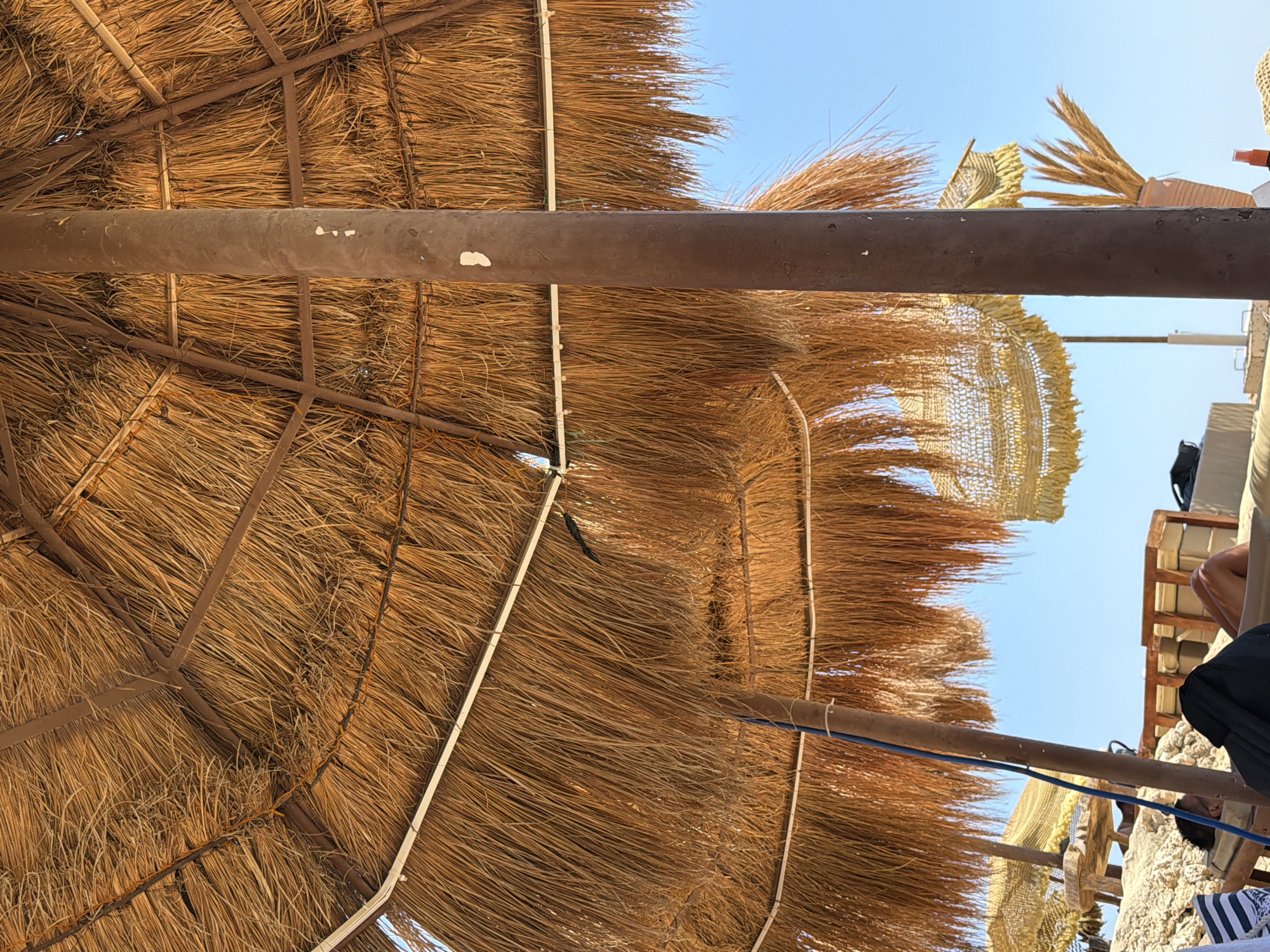 Underneath a large thatched umbrella, a person rests on a lounge chair on a beach in Sharm El Sheikh, Egypt. The umbrella, made of dried palm fronds supported by a metal frame, casts a shadow over the sandy area. In the background, another smaller thatched umbrella with decorative trim and a potted plant with dried reeds are visible against a clear blue sky. The scene depicts a typical beach setting, suggesting a relaxed atmosphere.