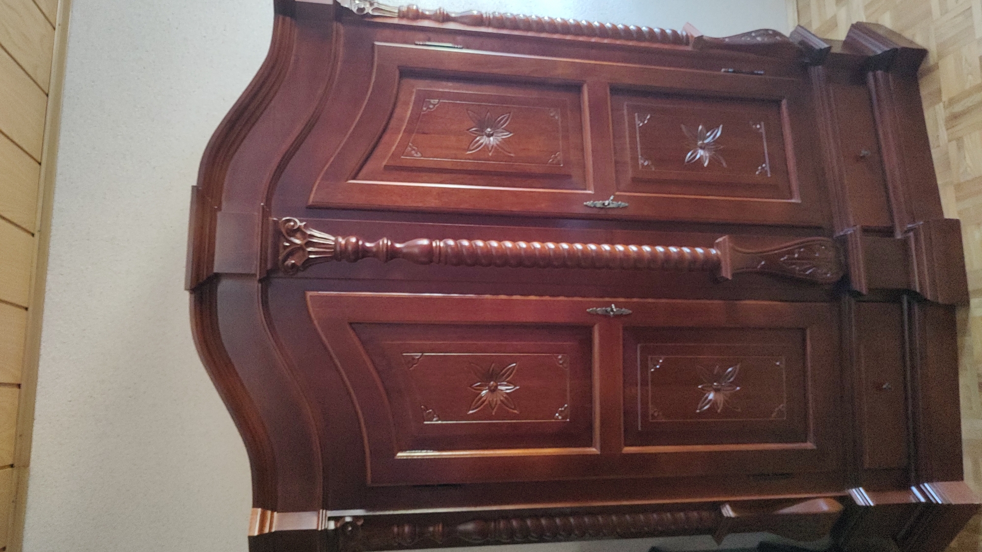 The image shows a dark wood wardrobe with two doors. The doors have decorative carvings of flowers. The wardrobe has ornate pillars on either side of the doors and at the corners. There are also drawers visible at the bottom of the wardrobe. The wardrobe is positioned against a light-colored wall, and a wooden ceiling is visible at the top of the image. The floor is made of light-colored wood planks. The image is taken from a frontal, slightly upward angle. There are no people or animals present in the image.