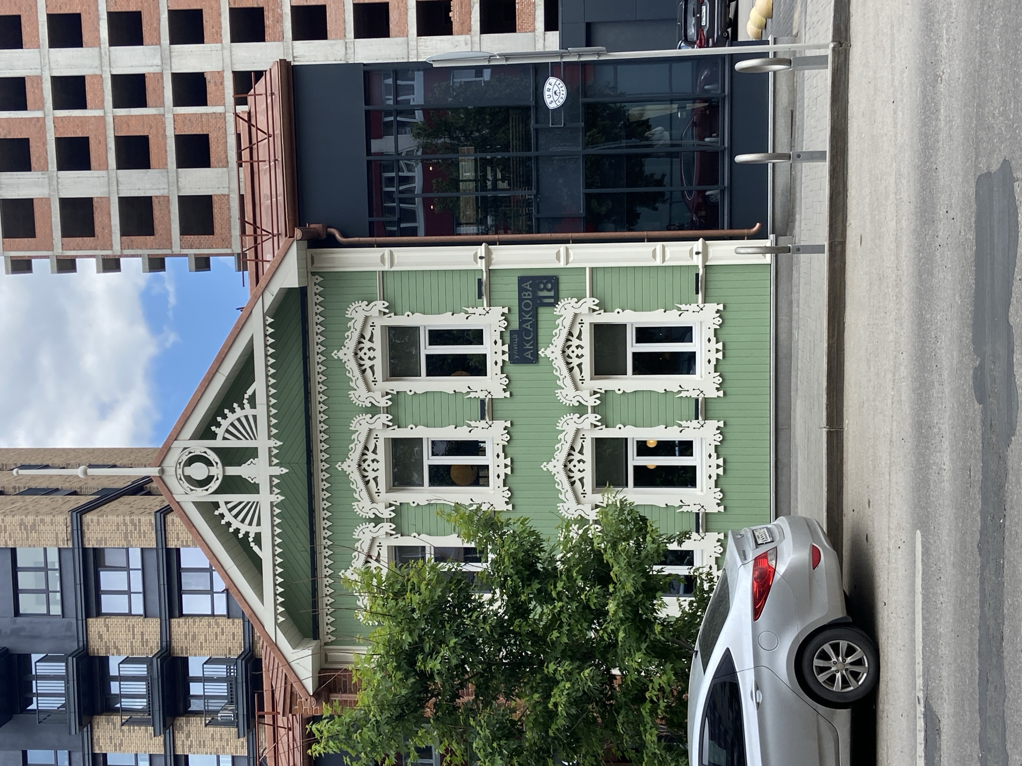 The image depicts a street scene in Ufa, Russia, in front of a building with a distinct green facade and intricate white trim around its windows. The building is a mixture of old and new architectural styles, with a traditional-looking wooden structure on the left and a modern, dark-glassed section to its right. Behind these structures, a multi-story brick building under construction is visible, along with a darker, modern apartment building. A silver sedan is parked on the street in front of the traditional building, and a tree with lush green foliage stands to the left. A street sign with Cyrillic text, "УЛ. АКСАКОВА 18", is mounted on the green building. The sky is partly cloudy. The scene appears to be a street in a city, with elements of residential and commercial architecture.