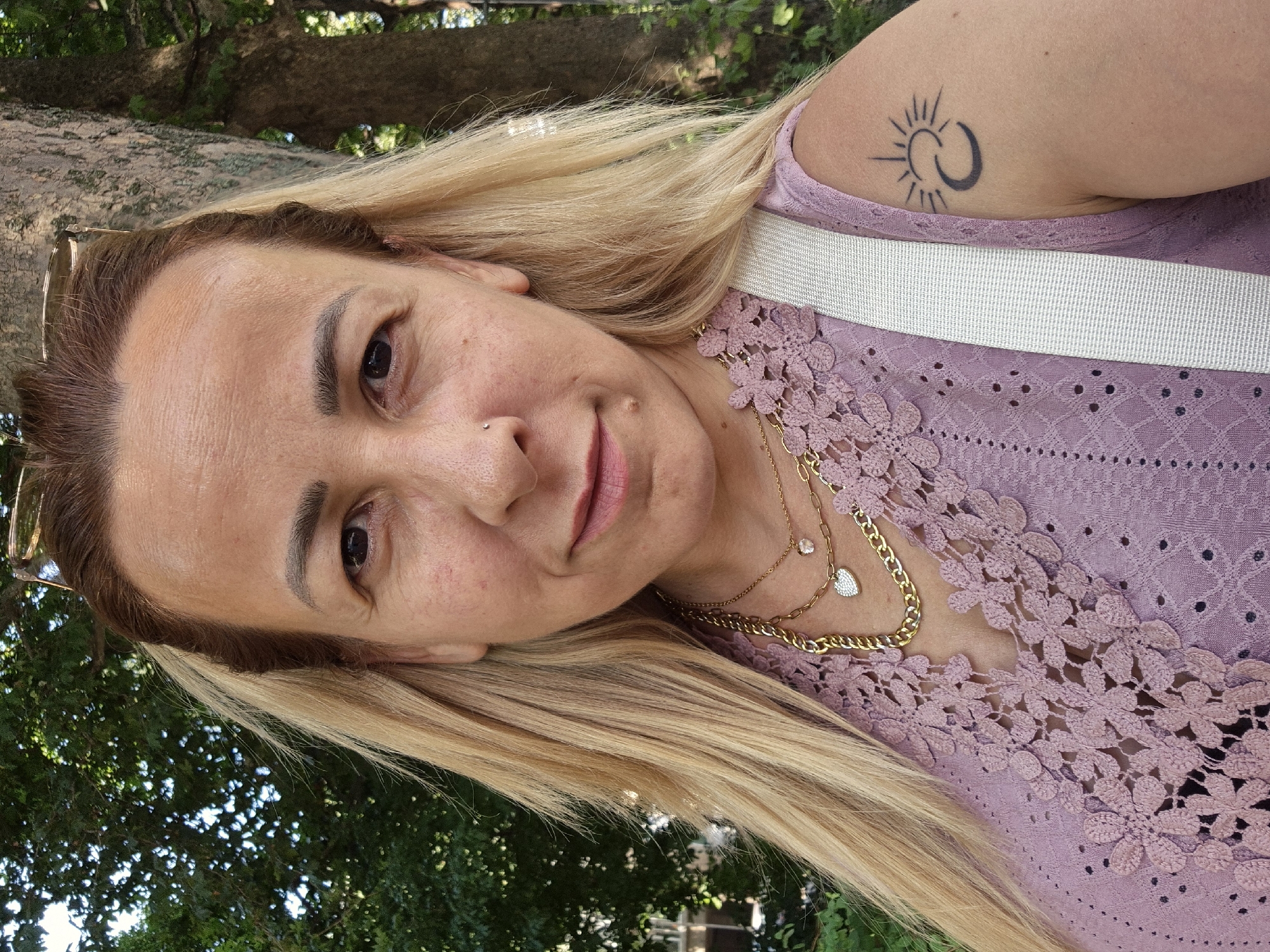 A close-up, eye-level, outdoor, selfie shows a light-skinned woman with long blonde hair styled in a center part. She is looking directly at the camera with a neutral expression. She is wearing a light purple, lacy blouse and several gold necklaces. A white strap from a bag or garment crosses her shoulder. A tattoo of a crescent moon and sun is visible on her upper right arm. The background consists of green foliage and a tree trunk. The lighting is natural and appears to be from a sunny day. The image is sharp and in focus.