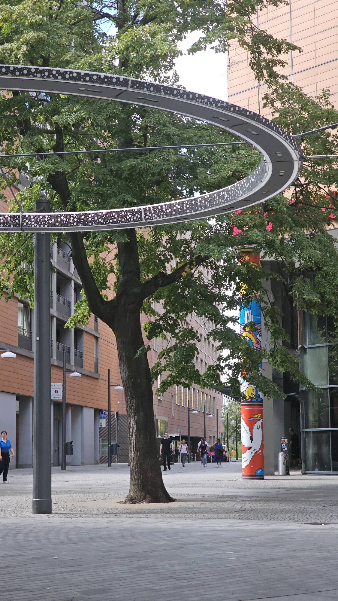 A mature tree with dense green foliage stands in the foreground, its trunk dark and textured. A large, circular metal structure with decorative LED lighting is suspended above and around the tree, casting a subtle glow. The structure appears to be a modern art installation or a decorative element of the urban landscape. In the background, modern brick buildings with multiple windows line a wide, paved pedestrian walkway. Several people are walking on the path, engaged in various activities such as strolling, carrying bags, and conversing. A tall, cylindrical column adorned with colorful illustrations, including a white bird-like figure, stands to the right of the tree. The environment suggests a public space in a European city, specifically Berlin, Germany, given the architectural style and the presence of a sign with German text, possibly indicating a public service or landmark. The overall scene depicts a vibrant urban setting with a blend of natural elements and contemporary design.