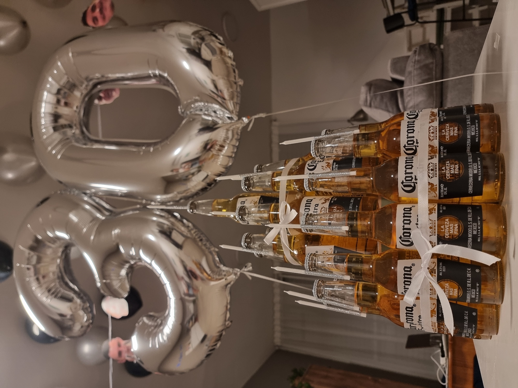 An indoor, close-up, eye-level shot captures a celebratory arrangement of Corona beer bottles and silver balloons forming the number "30". The balloons, positioned above the bottles, are large and reflective, with a distinct "3" on the left and a "0" on the right. Embedded within the balloons are printed images of a man's face, appearing multiple times. The beer bottles are stacked in tiers, with white ribbon tied around the necks of some. Each bottle has a Corona Extra label. White candles are inserted into the necks of several bottles. The background is blurred, showing hints of furniture, possibly a sofa, and a chair, suggesting a domestic or event space. The overall scene is lit by ambient indoor lighting, creating reflections on the metallic balloons and glass bottles. There is no indication of the city of Ramat Gan, Israel in the image.