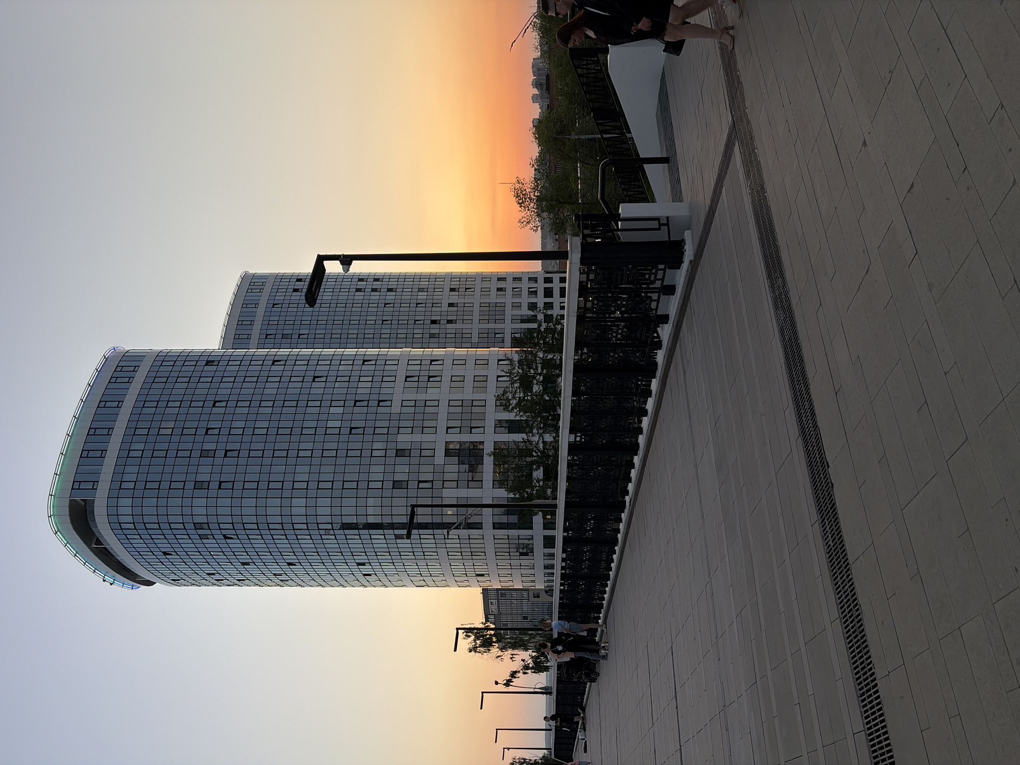The image is a street-level view in Izhevsk, Russia, showcasing modern architecture under a twilight or sunset sky. Two tall, curved buildings with grid-like windows dominate the left side of the frame. Pedestrians are visible walking on a paved walkway. The sky fades from orange near the horizon to lighter shades above. Street lamps line the walkway. A black metal railing separates the walkway from the buildings. The pavement consists of large, rectangular slabs, with a drainage grate running along the walkway.