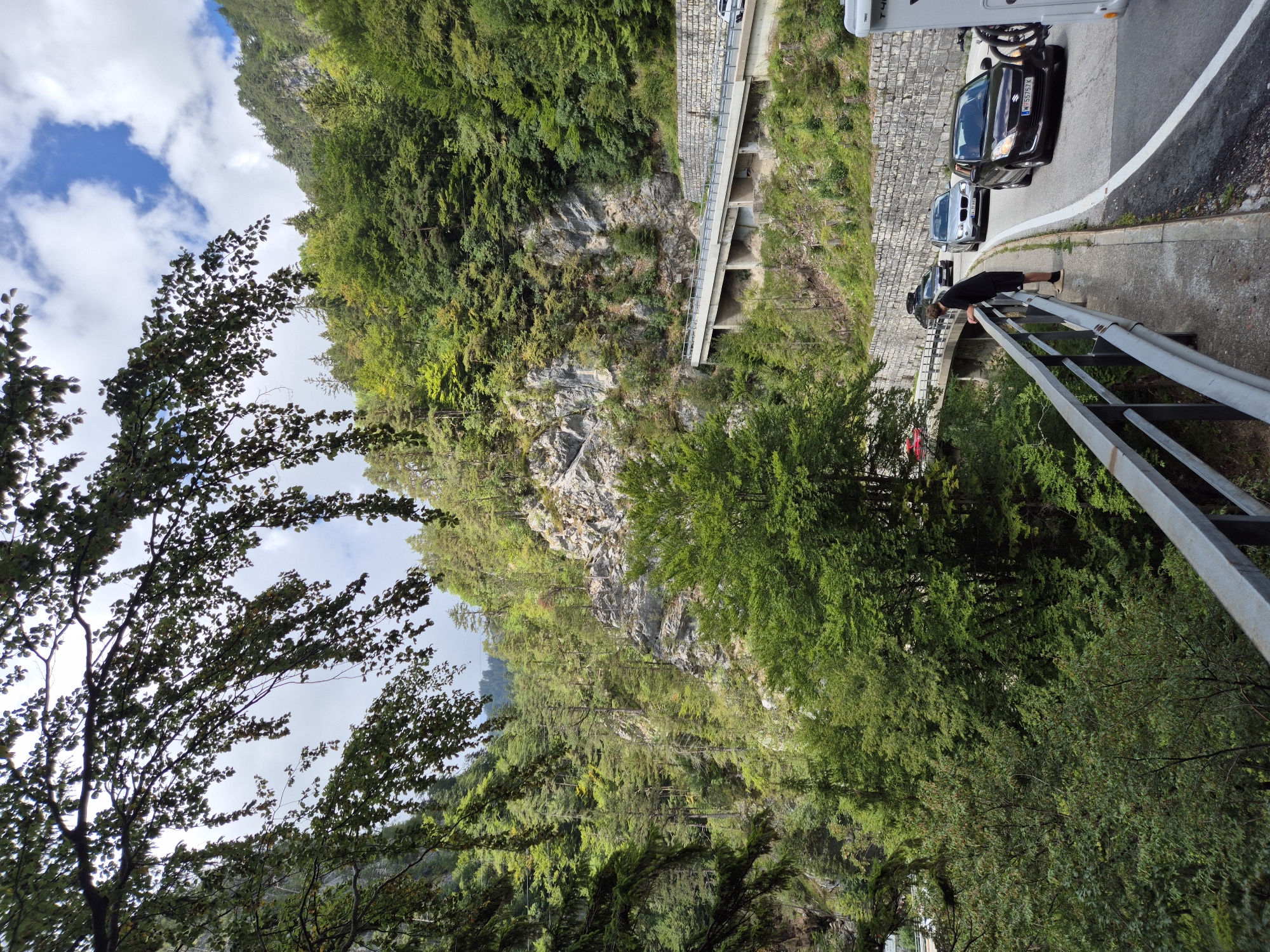 In the city of Ferlach, Austria, a person stands on a pedestrian walkway overlooking a road with several cars. The walkway is bordered by a metal railing. The road curves around a steep, tree-covered mountainside. A white camper van is visible on the right side of the road, followed by a dark brown Suzuki Swift with a license plate that reads "W 55757 K." Other cars are also present on the road, including a silver sedan and a black SUV. The mountains are covered in lush green trees, and the sky is partly cloudy. The scene appears to be a scenic view along a mountain road, possibly a tourist destination.