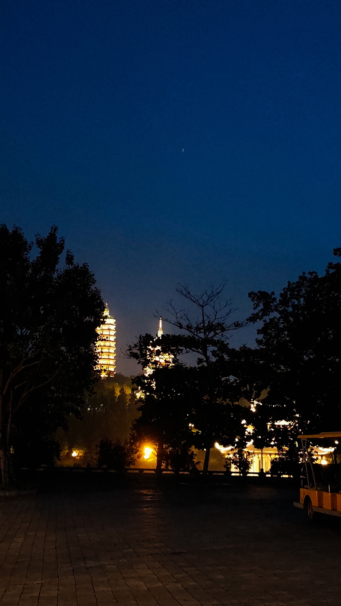 The image captures a night view of pagodas in Tây Hoa Lư Ward, Vietnam. Two illuminated pagodas are visible in the background, towering over lush greenery. The foreground features a paved walkway and silhouetted trees. A small, yellow electric vehicle is parked on the right side of the image. The sky is a deep, clear blue, with a faint celestial object visible. The lighting suggests a tranquil, possibly spiritual, atmosphere.