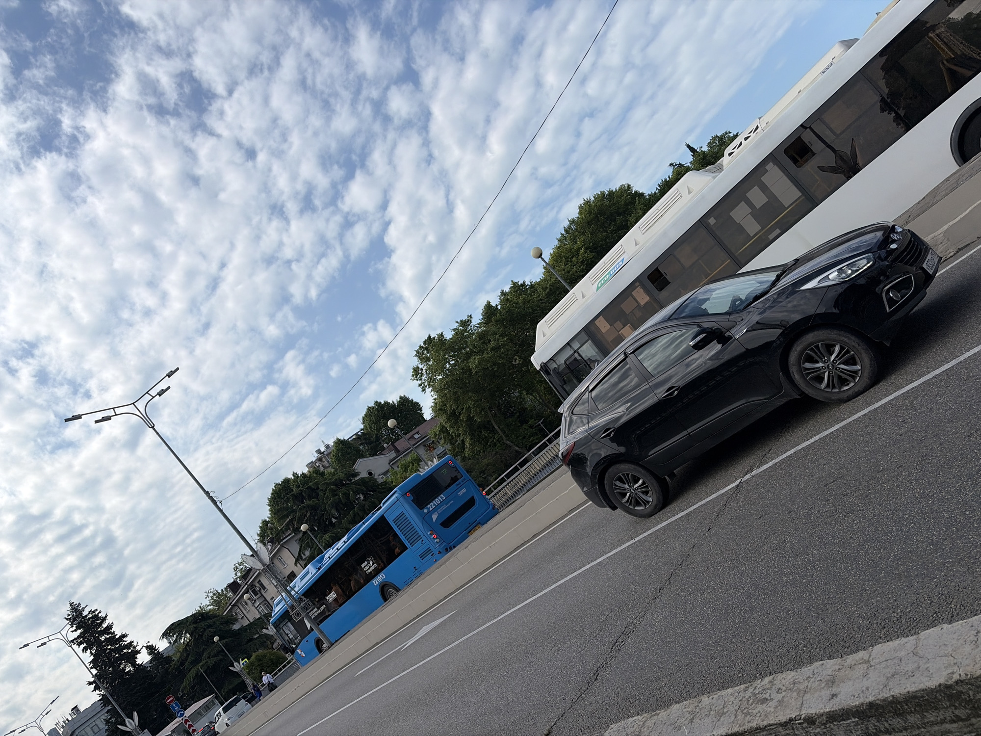 This is a tilted, eye-level, outdoor, daytime shot of vehicles on a road in Sochi, Russia. In the foreground, a black SUV is parked on the right side of the frame. To the left of the SUV, a blue bus is traveling on the road. Behind the blue bus, a white bus is also traveling. The road is paved and has white lane markings. The background consists of lush green trees and buildings. The sky is blue with scattered white clouds. There are several street lights visible, including a tall, modern one with multiple lamps. The image is dominated by the vehicles and the road, with the natural and urban elements serving as a backdrop.