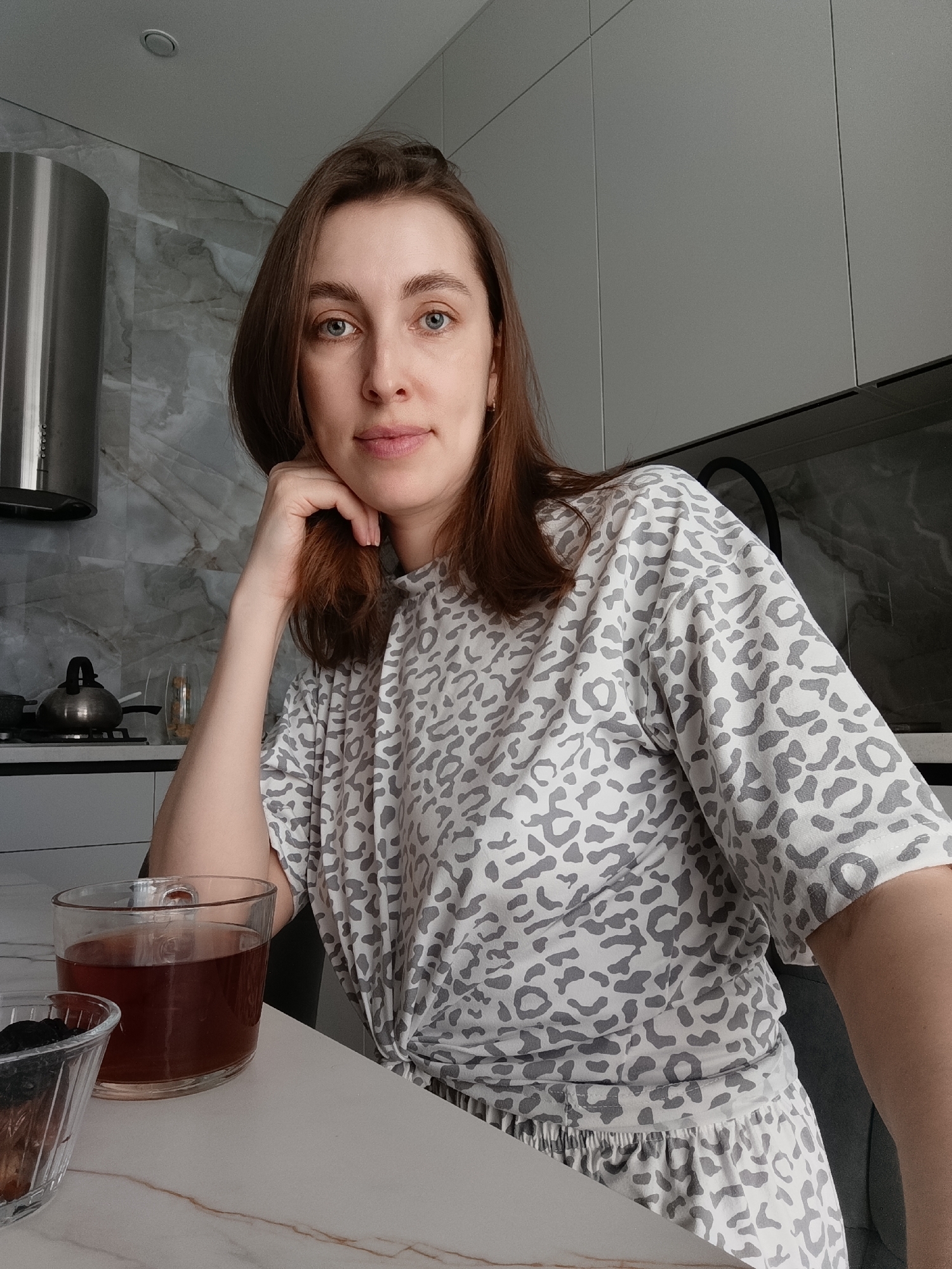 An adult female individual is depicted in a modern kitchen setting in the city of Chaikovsky, Russia. She has medium-length brown hair and blue eyes, and is looking directly at the viewer with a neutral expression. Her left hand is positioned under her jawline. She is wearing a light-colored shirt with a grey leopard print pattern, and matching bottoms are partially visible. On a white countertop with subtle brown veining in the lower left, a clear glass mug filled with a dark reddish-brown liquid, consistent with tea, is present alongside a small clear fluted bowl containing dark dried fruit. The background displays a kitchen equipped with a stainless steel cylindrical range hood, a black stovetop with a silver kettle, and a grey marble-effect tiled backsplash. Grey flat-panel overhead cabinets are visible to the right, and a recessed ceiling light fixture is present.