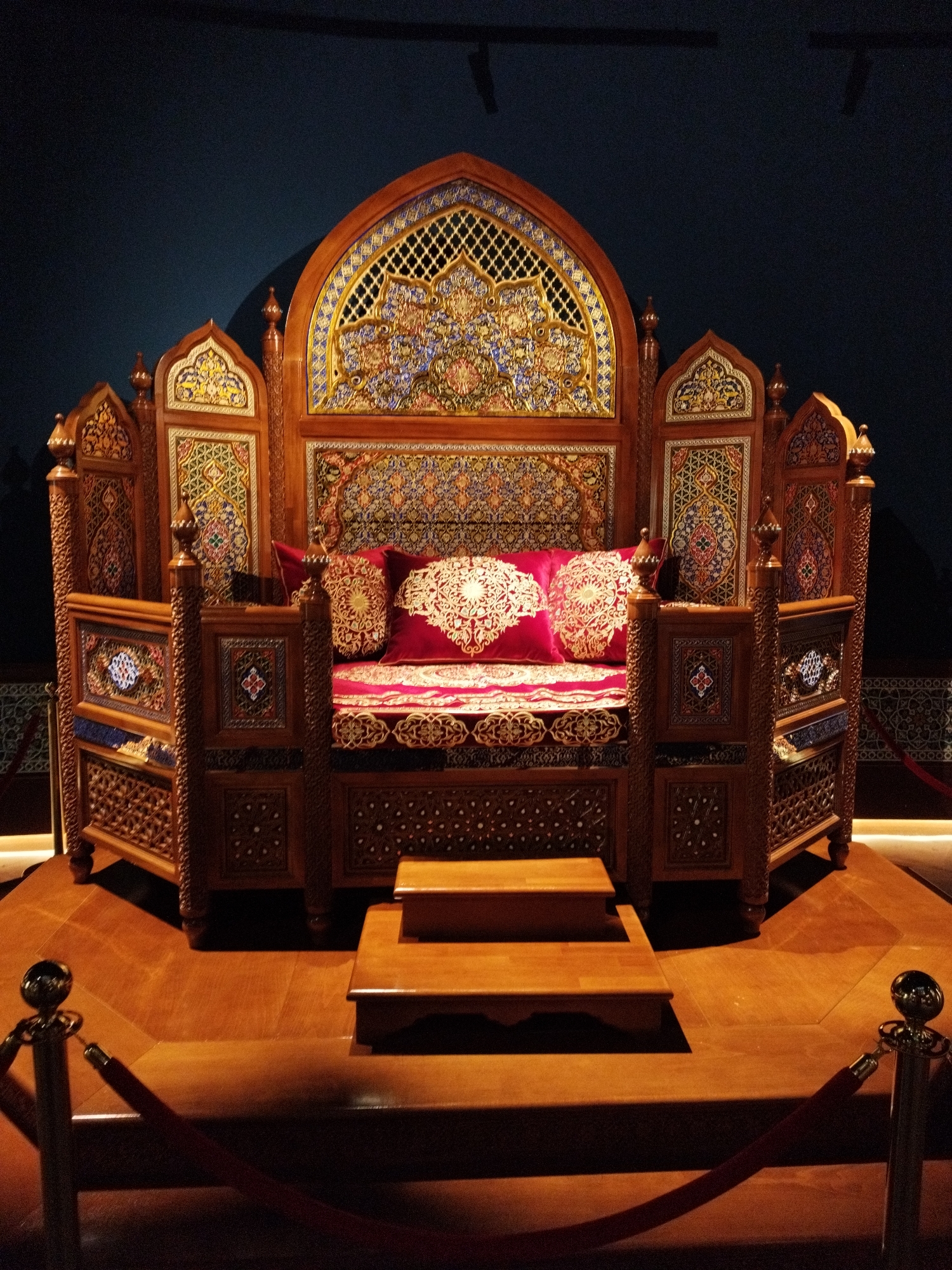 An ornate wooden throne or daybed, elaborately carved and inlaid with colorful mosaic tiles, is displayed on a wooden platform in a dimly lit room. The throne features a high, arched headboard with intricate geometric patterns and a central panel depicting a floral motif. The side panels and the base of the throne are also adorned with detailed carvings and mosaic inlays. The seating area is covered with a red cushioned mattress and four red throw pillows with gold circular patterns. In front of the throne, there are two wooden steps, also stained a rich brown. The background is a dark, solid wall. Ropes and stanchions are visible in the foreground, suggesting the object is part of a museum exhibit. The lighting creates strong shadows, emphasizing the rich textures and details of the throne.