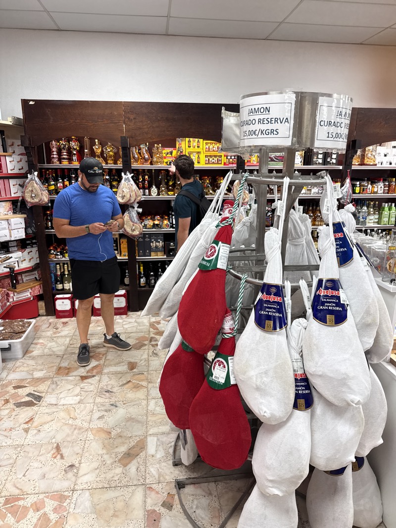 The image captures the interior of a store likely specializing in Spanish delicacies, specifically showcasing cured ham ("Jamon"). A rotating display in the foreground features several hams, some covered in white protective wrappings and others in red. Labels indicate the ham is "Jamon Curado Reserva" and "Jamon Gran Reserva," with a price of 15.00€/KGRS. Prominently displayed on the ham packaging is the brand name "Quejusa" and "Salamanca," a region known for its cured ham. Two men are visible: one wearing a blue shirt, black shorts, and a baseball cap, looking at his mobile phone, and another man with a backpack inspecting items on a shelf. Shelves in the background are stocked with a variety of items, including bottles of wine and other gourmet products. The store has a tiled floor in a mosaic pattern. The details "Salamanca" and price tags suggest the image was taken in Fuentes de Oñoro, Spain.