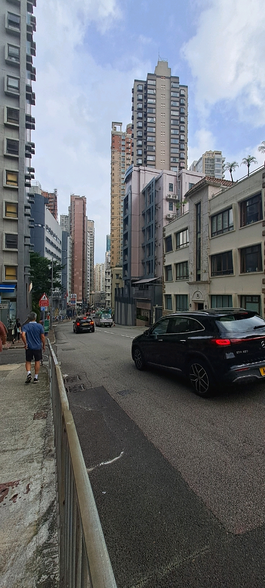 A street scene in a dense urban environment with tall buildings lining both sides of the road. The perspective is from a sidewalk on the left, looking down the street. A man in a blue polo shirt and black shorts is walking away from the camera on the sidewalk. A black SUV is driving on the right side of the road, followed by other vehicles further down. The buildings are a mix of modern and older architecture, with many windows and balconies visible. The sky is cloudy. The environment appears to be a commercial or residential area within Hong Kong.