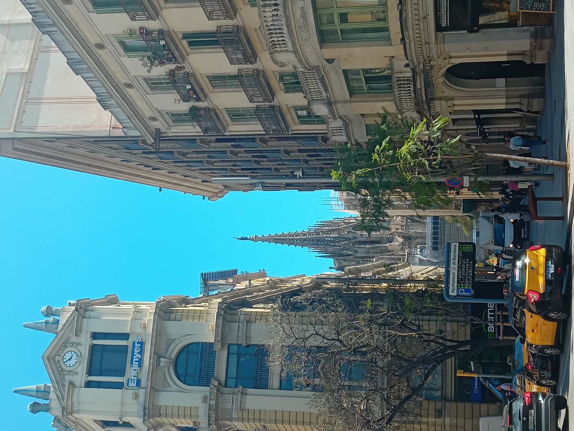 The image shows a street in Barcelona, Spain. The buildings on either side of the street have architectural details like balconies and ornate windows. The "Enginyers" building is visible on one side with a clock. The spire of the Barcelona Cathedral (also known as Catedral de la Santa Creu i Santa Eulàlia) is visible in the background.
There are trees along the street. The street has traffic, including cars, taxis, and pedestrians. Street signs and advertising are also present.