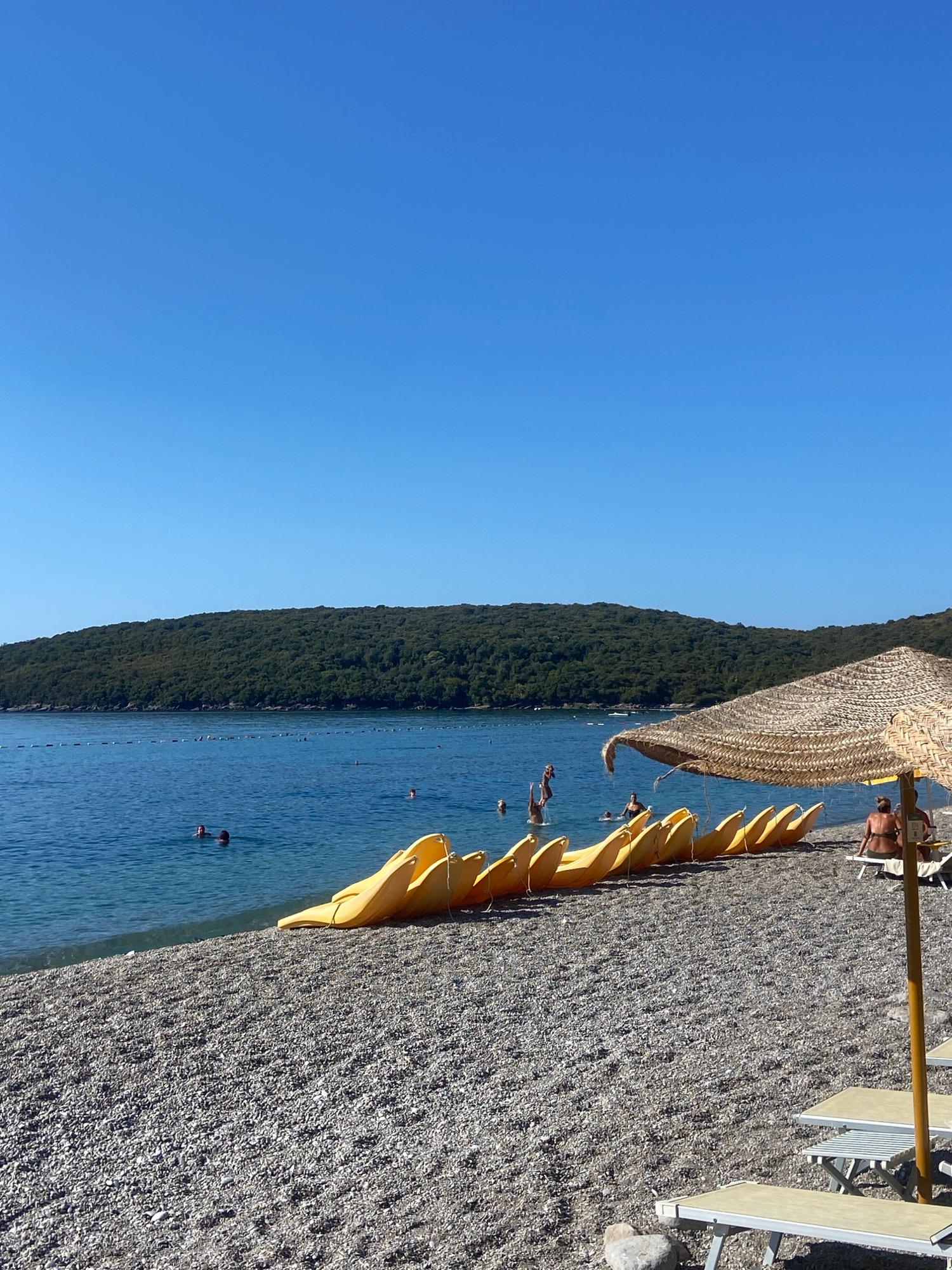 The image depicts a sunny day at a pebble beach in Prijevor, Montenegro. In the foreground, a row of yellow banana boats is neatly arranged on the shore, with a large straw umbrella providing shade over two lounge chairs. Further back, the clear blue sea is dotted with several people swimming. A floating rope barrier marks a designated swimming area. The background features a lush, green hillside, indicating a natural, vegetated landscape. The sky is a vibrant, cloudless blue, characteristic of a warm, summer day.
