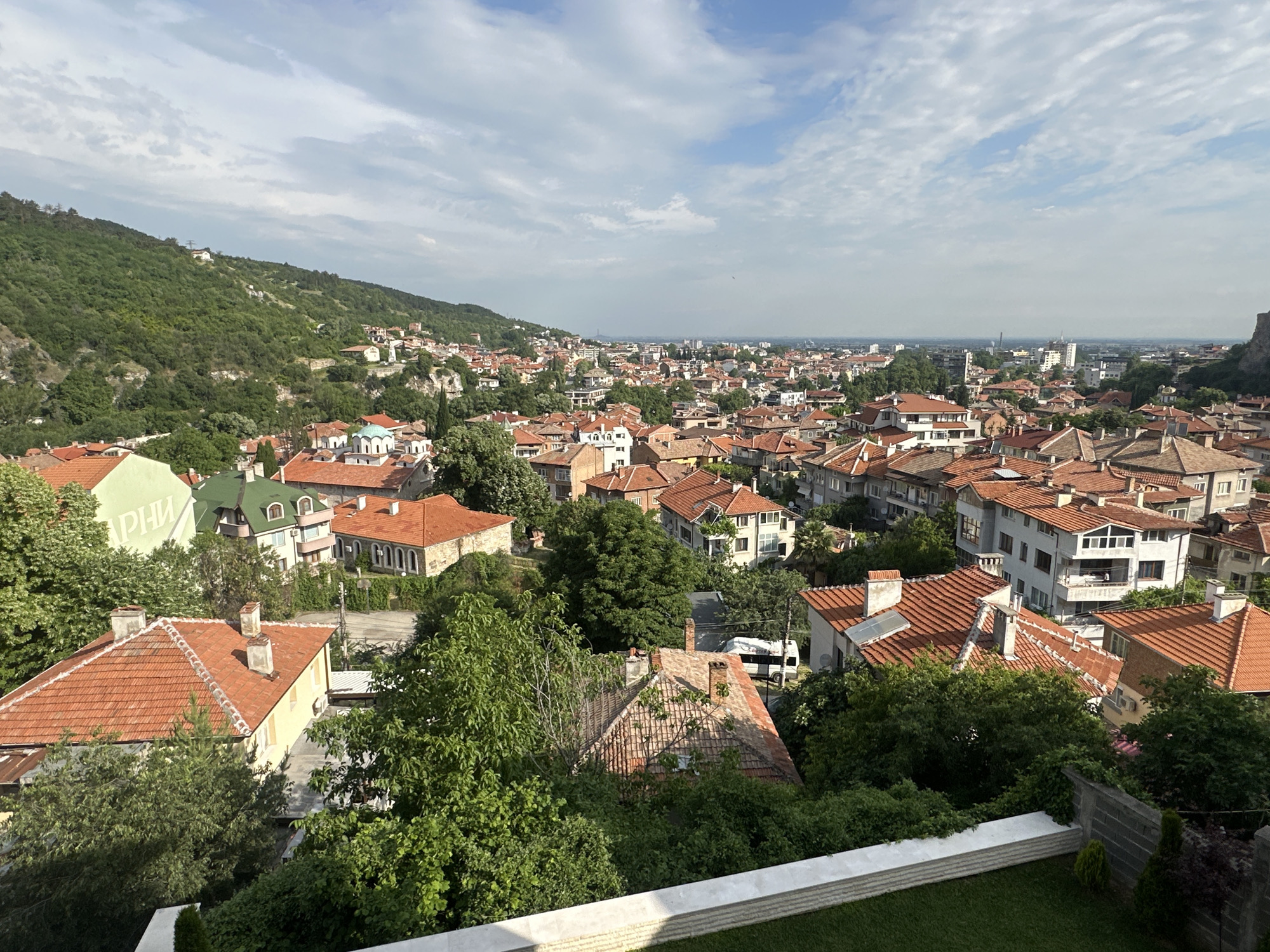 This elevated panoramic view captures the town of Asenovgrad, Bulgaria, nestled amongst lush, green hills under a bright, partly cloudy sky. The townscape is characterized by a dense array of buildings, predominantly houses featuring distinctive red and orange terracotta tiled roofs, indicative of traditional Balkan architecture. Abundant green trees and shrubs are interspersed throughout the urban fabric and thickly cover the surrounding hilly terrain.
In the immediate foreground, a white, low wall and a strip of green grass suggest the photograph was taken from a balcony or terrace. The town extends far into the distance, with some taller, more modern buildings visible in the background. In the mid-ground, a building with a light green dome roof stands out, possibly a church. Sparse white structures are also visible on the distant hillsides.
The sky is a mix of clear blue and scattered white, wispy clouds, suggesting a pleasant daytime, likely morning or late afternoon. No human activity is overtly visible, contributing to a tranquil and serene atmosphere. On a light green building towards the mid-left, partially visible Cyrillic text can be discerned. The scene conveys the picturesque charm of a historic town harmoniously integrated with its natural, hilly environment.