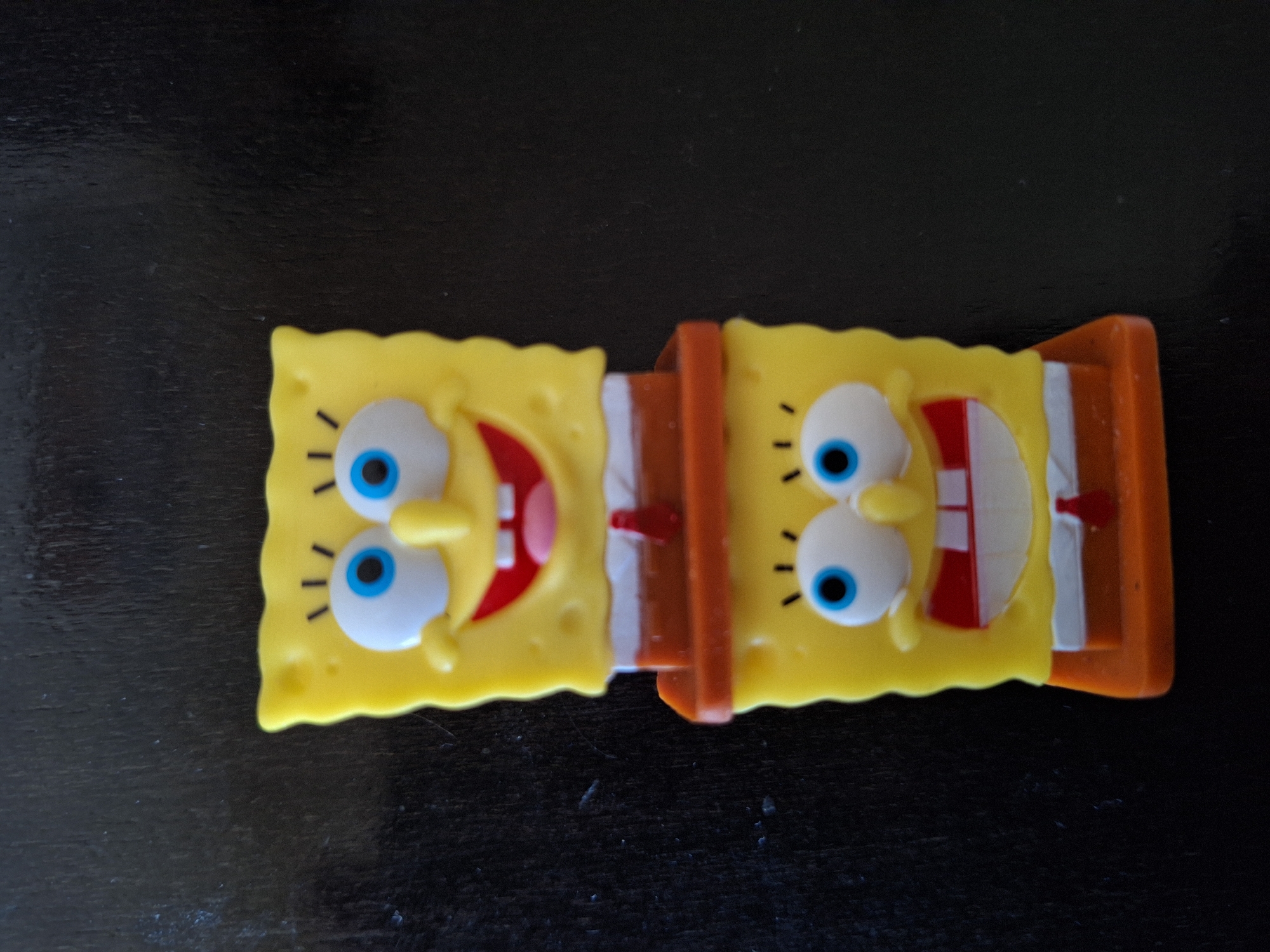 The image shows two Spongebob Squarepants candy dispensers connected by an orange plastic piece, lying horizontally on a dark, textured surface. The dispensers feature Spongebob's character design: yellow, porous faces with protruding blue eyes, noses, and open mouths showing teeth and red tongues. The base of the dispenser is orange and white, resembling his pants. The scene is indoors on what appears to be a dark wood or plastic surface. Location: Unknown, Unknown.