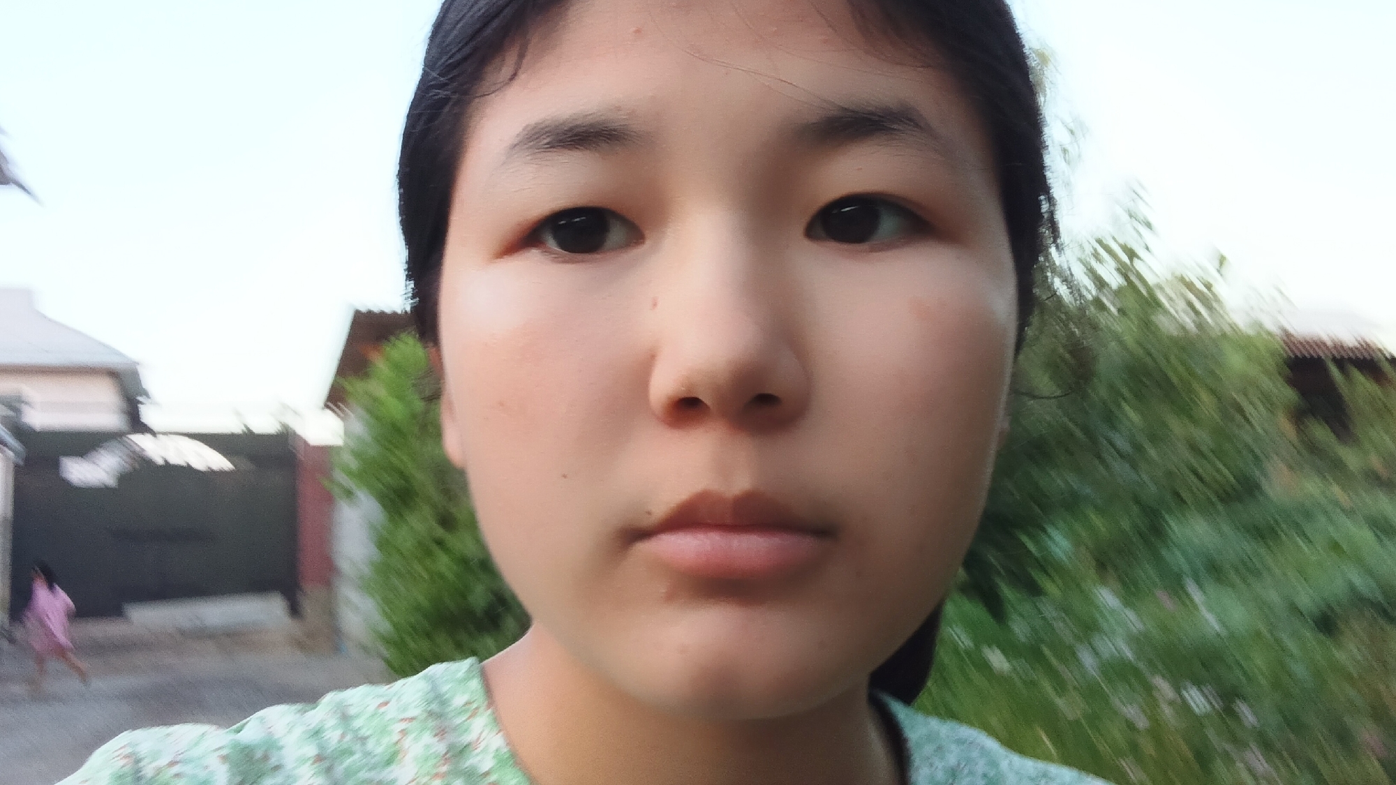 This is a close-up, selfie-style portrait of a young East Asian individual, likely a female, looking directly at the camera. The subject is positioned in the foreground, with their face dominating the frame. They have dark hair, dark eyes, and light skin with a slight blush on their cheeks. Their lips are slightly parted. They are wearing a green shirt with a floral or leaf-like pattern.
The background is out of focus, but suggests an outdoor setting. To the left, there are hints of buildings and a dark gate, and a person in a pink dress can be seen walking in the distance. To the right, lush green foliage and trees are visible, with hints of a building roof behind them. The lighting suggests it is daytime, possibly late afternoon or early evening due to the soft natural light and the slightly hazy sky. The overall impression is casual and informal, possibly taken in a residential area given the buildings and yard. No text is visible in the image.