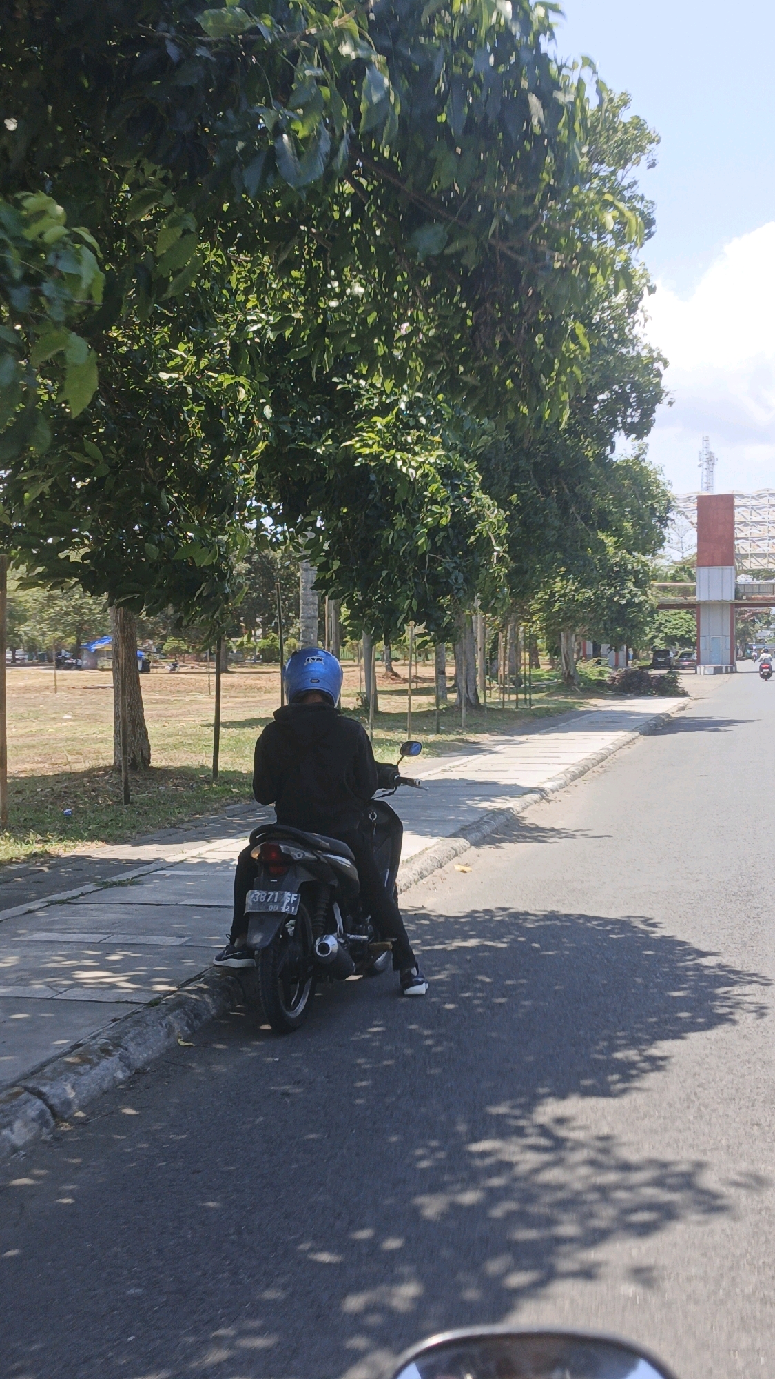 A person wearing a blue helmet and a black hoodie sits on a black motorcycle parked on the side of a paved road in Pangandaran, Indonesia. The motorcycle's license plate is visible. The road is bordered by a sidewalk and a line of trees with dense green foliage. Beyond the trees, there is a grassy field with some scattered smaller trees and structures, including what appears to be a blue tarped shelter and a distant building with red and white accents. The sky is clear and blue, with a few scattered white clouds. Sunlight casts dappled shadows on the road from the trees.