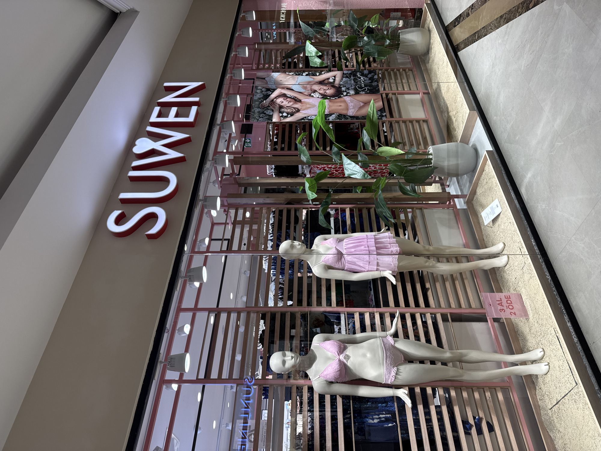 This image shows the storefront of a lingerie shop named SUWEN, located in Bodrum, Turkey. The shop features a modern facade with a large glass window displaying two mannequins wearing pink lingerie. The mannequins are positioned prominently in the foreground, one on the left and the other on the right. Behind the mannequins, shelves are stocked with clothing items. A promotional sign in pink, reading "3 AL 2 ÖDE" (which translates to "Buy 3, Pay for 2"), is placed on the floor in front of the left mannequin. The shop's name, "SUWEN," is illuminated in red and white capital letters above the window. Potted plants are strategically placed to enhance the store's visual appeal. The interior of the store is visible through the glass, revealing racks of garments and additional display elements. The floor outside the store is tiled in a light gray stone pattern with decorative marble inlays. The overall environment suggests a commercial setting within a mall or shopping district.