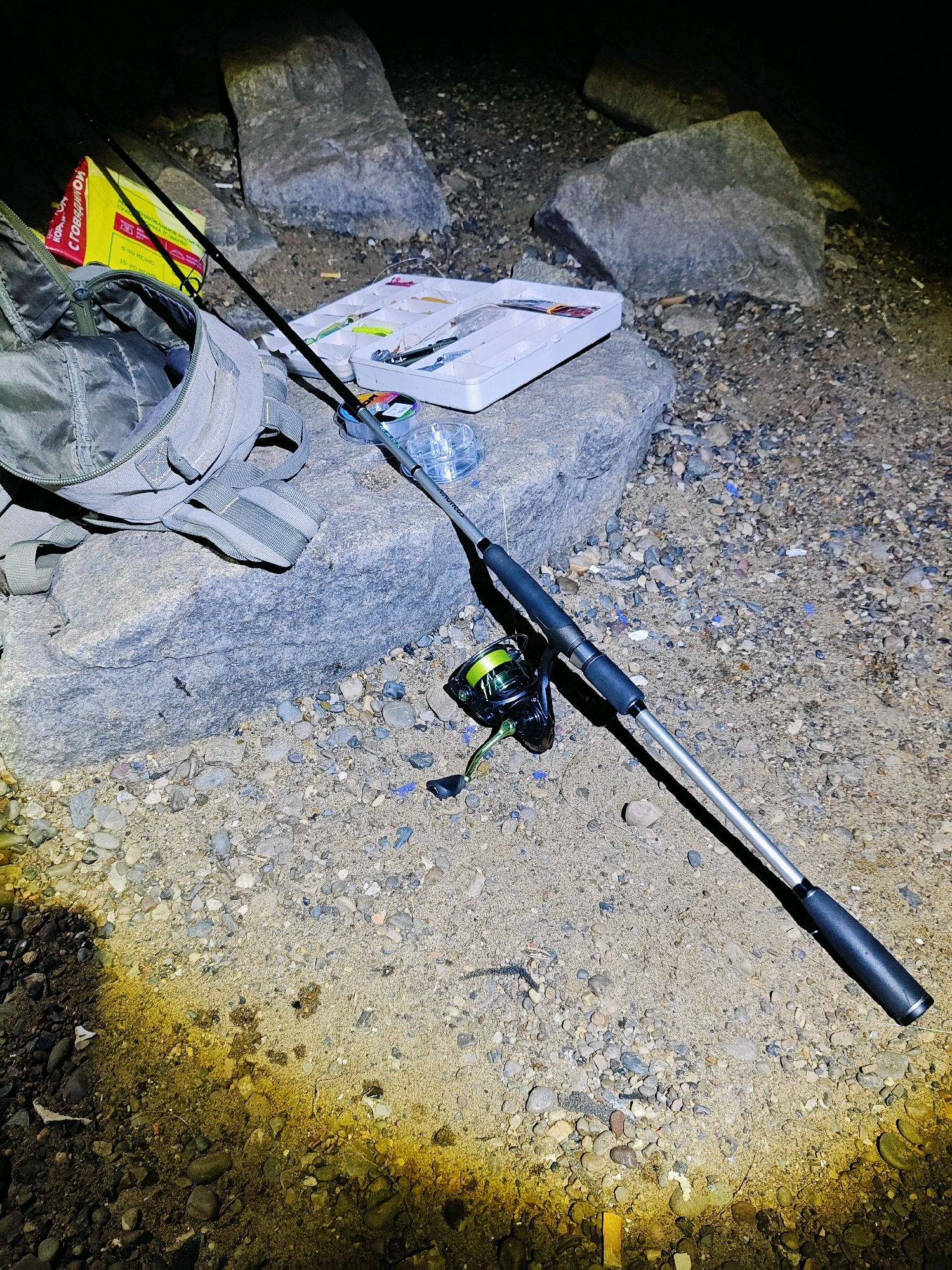 A fishing rod, reel, tackle box, and a backpack are laid out on a rocky shore. The scene is illuminated by a spotlight, casting long shadows. The tackle box is open, revealing fishing lures and other accessories. The backpack is partially open, showing its contents. The ground is covered in small pebbles and sand. The location is near the Kama River in Chaikovsky, Russia.