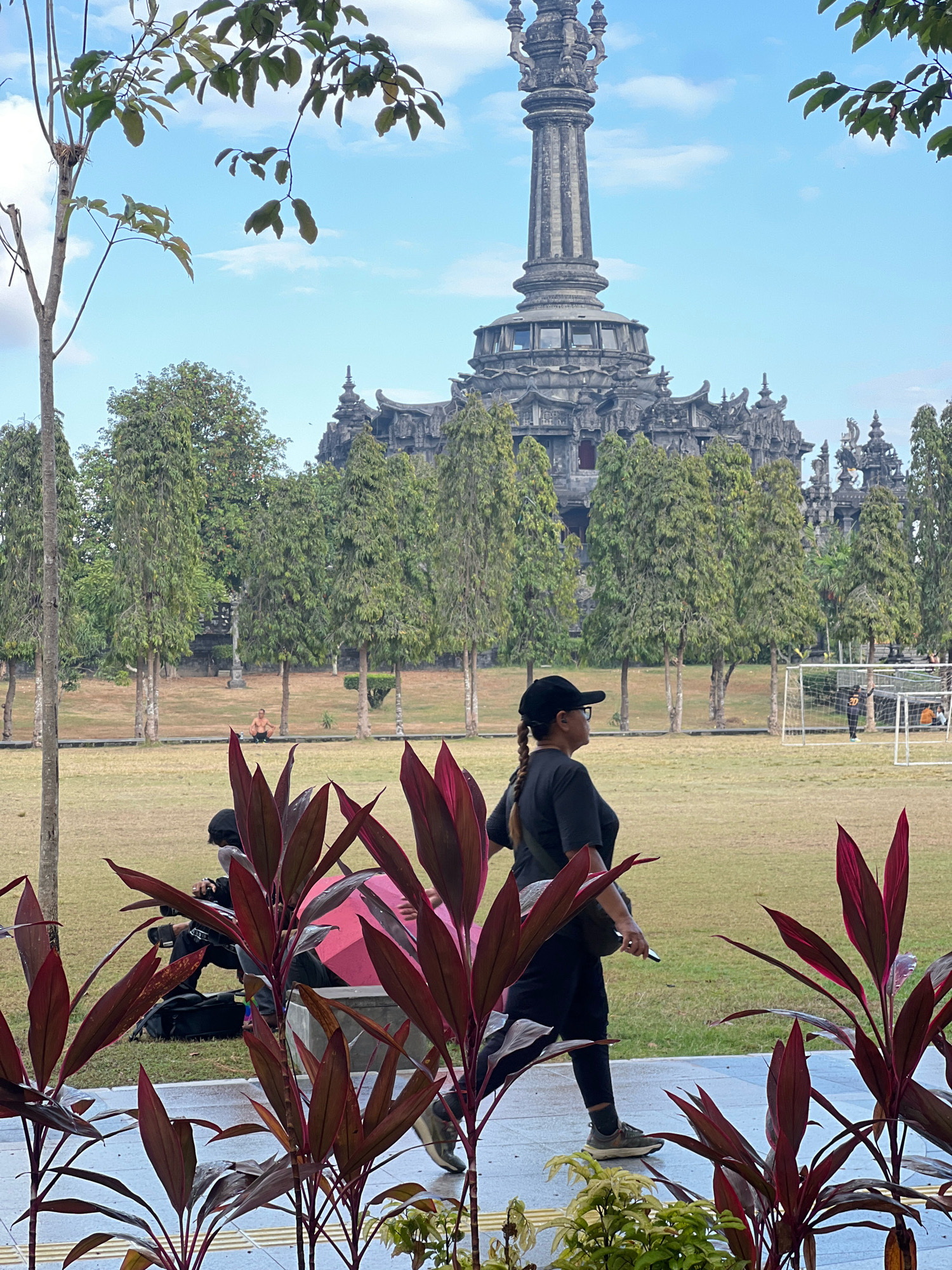 The image captures a daytime scene at the Bajra Sandhi Monument in Denpasar, Bali, Indonesia. In the foreground, a woman wearing a black cap, t-shirt, and pants walks on a paved path, holding a phone in her right hand. She is seen from the side, facing left, with her hair in a braid. To her left, a motorcycle is parked. In the middle ground, a man is seated on the grass. A soccer field with a goalpost is visible on the right side of the image, with a few people in the distance. The Bajra Sandhi Monument, a large, grey, ornate structure, dominates the background, with trees lining the manicured lawn. The sky is blue with scattered clouds, and the overall lighting suggests it is late afternoon. Red-leafed plants are in focus in the immediate foreground, framing the scene.