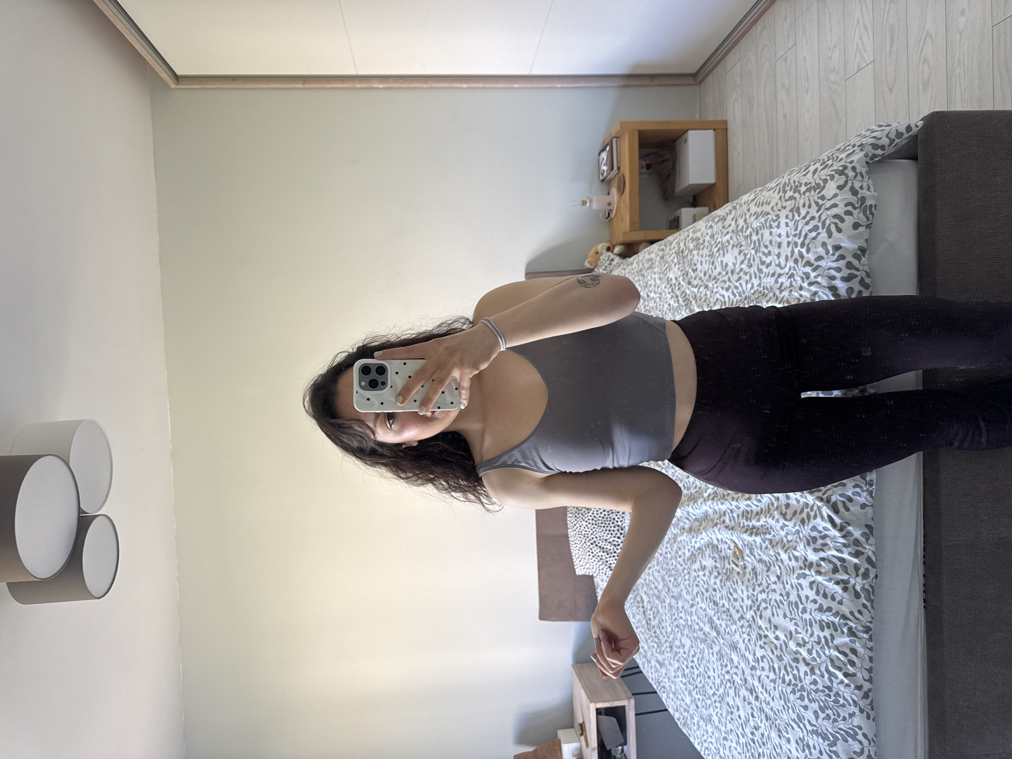 A young woman is taking a selfie in front of a mirror. She is wearing a gray tank top and dark leggings. She is holding a white cell phone with a speckled case. She has dark, curly hair. The mirror reflects a bedroom scene: a bed with a patterned comforter, a bedside table with decorative items, and a wall with a modern light fixture. The location cannot be determined to be in Burgas, Bulgaria from the image.