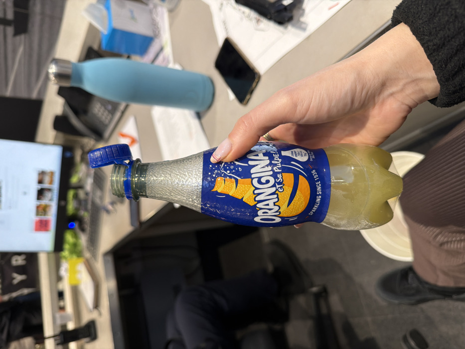 A hand holds an open bottle of Orangina soda. The bottle's label is blue with the Orangina logo. Inside the bottle, the liquid is a yellow-orange color. A blue water bottle sits nearby. A cell phone, paperwork, and part of a computer are visible in the background. The person holding the bottle wears black clothing. The person's legs are also visible, clad in brown pants and black shoes. A white container is visible to the right of the drink.