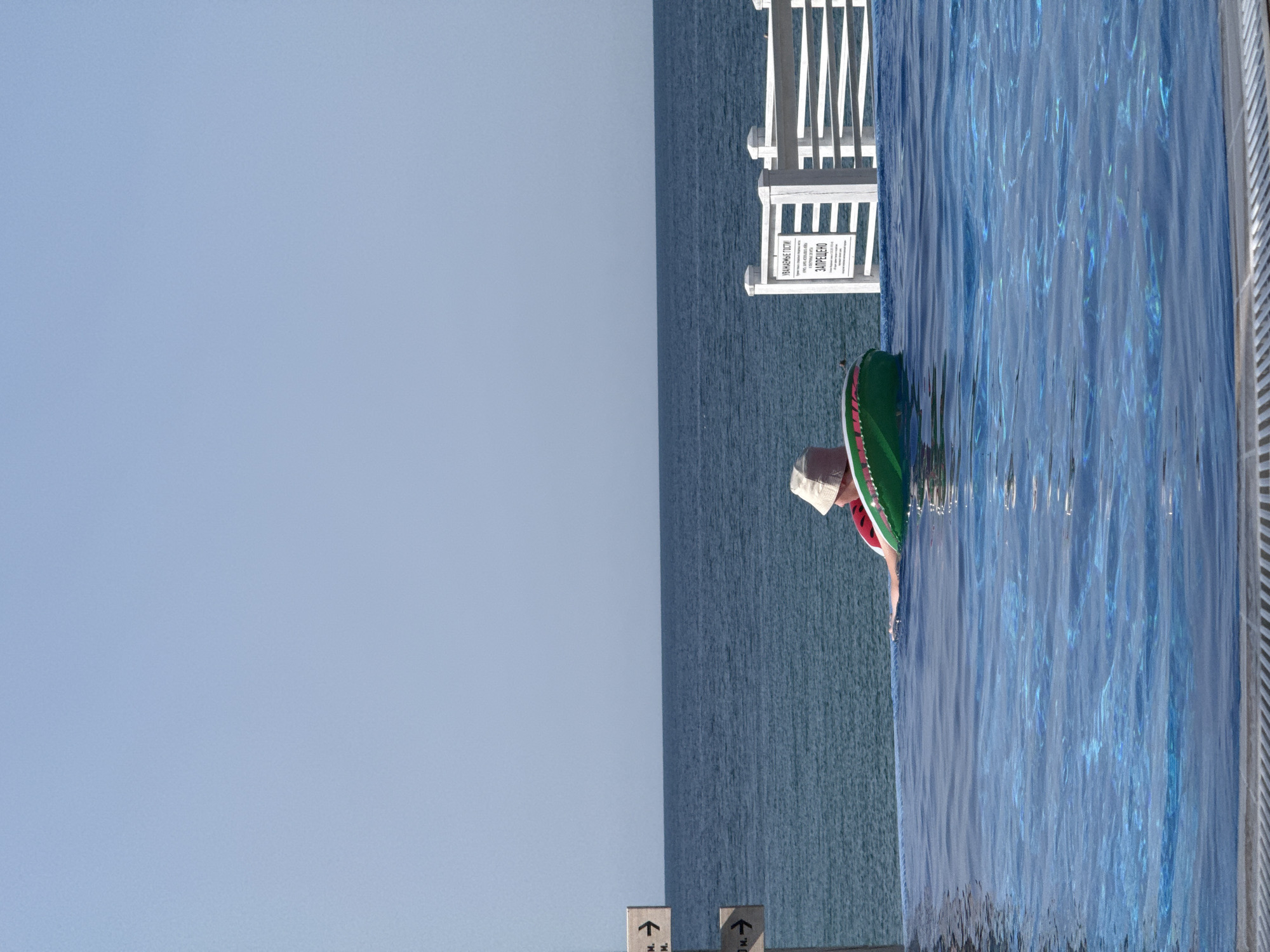 A person wearing a hat and a red shirt floats in an infinity pool on a clear day, looking out at the ocean. The person is in a green and pink inflatable ring. There are signs in the background, one pointing to the left with text that appears to be in Cyrillic, and another white sign with text in Cyrillic that reads "УВАЖАЕМЫЕ ГОСТИ! ЗАПРЕЩЕНО" which translates to "DEAR GUESTS! PROHIBITED". A white railing separates the pool from the ocean. The water in the pool is blue and reflects the sunlight.