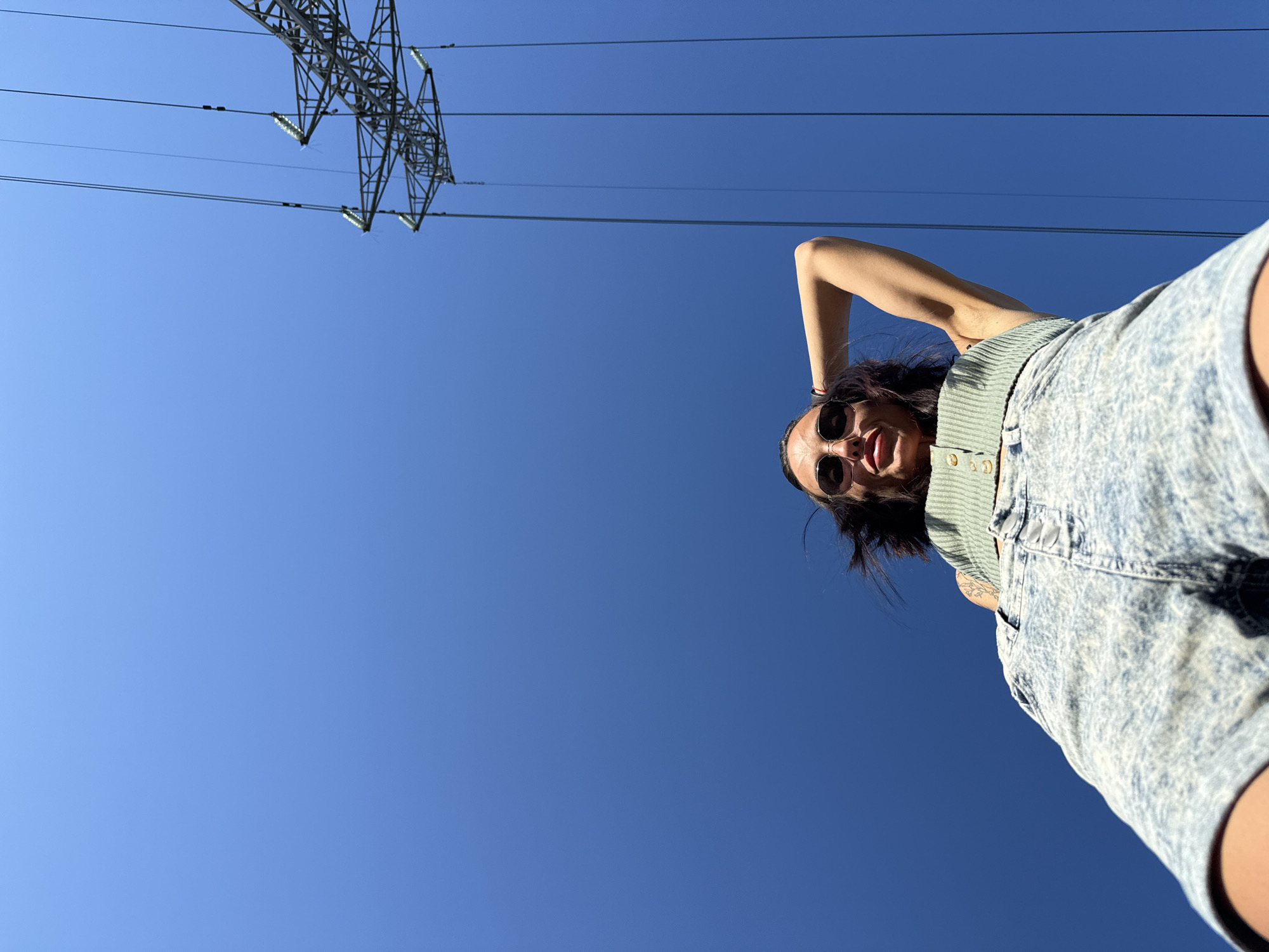 A woman wearing sunglasses, a green top, and acid-wash denim shorts is viewed from a low angle against a clear blue sky. In the upper left corner of the frame, a power line tower is visible with power lines stretching across the sky. The woman is positioned towards the right side of the image, with one arm raised behind her head. The specific location in Pleven, Bulgaria cannot be determined from the image.