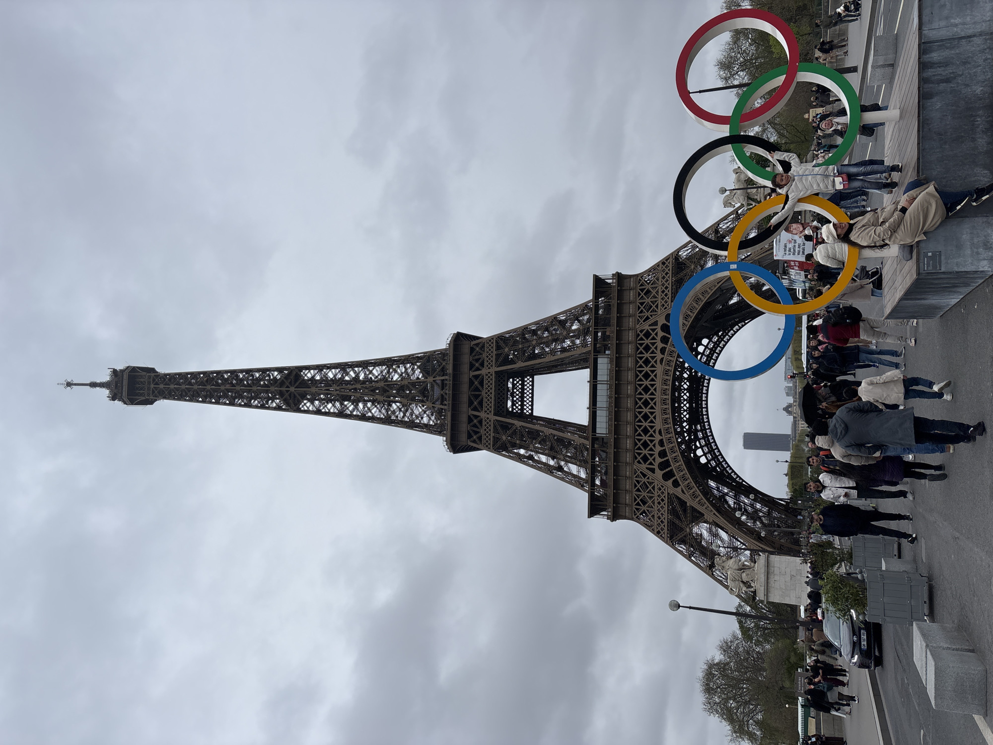 The image captures the Eiffel Tower in Paris, France, positioned under a cloudy sky. Five Olympic rings are prominently displayed near the base of the tower, with people interacting with the display. Several people are standing and seated near the rings, appearing to pose for photos. A group of tourists is visible in the background, observing the scene. There are trees and street furniture along the roadway. Cars are also present on the street.
