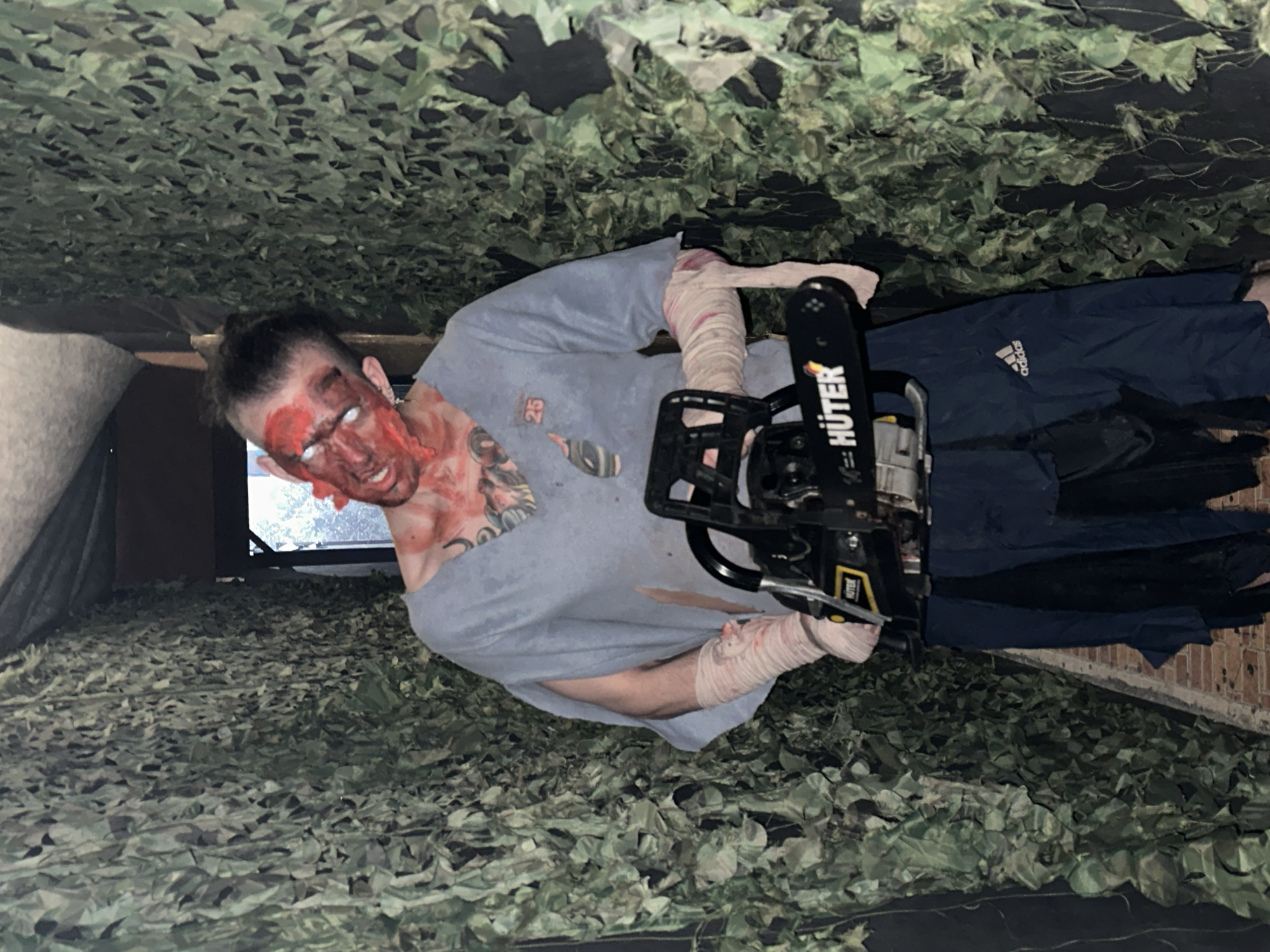 A person with a bloodied face and white contact lenses, possibly a zombie costume, is holding a chainsaw. The person is wearing a gray t-shirt, bandages on their arm, and blue Adidas pants. The scene is set in a camouflaged environment, possibly an indoor or outdoor location with netting or camouflage material. The location is not Saratov, Russia, as that information is not provided in the image.